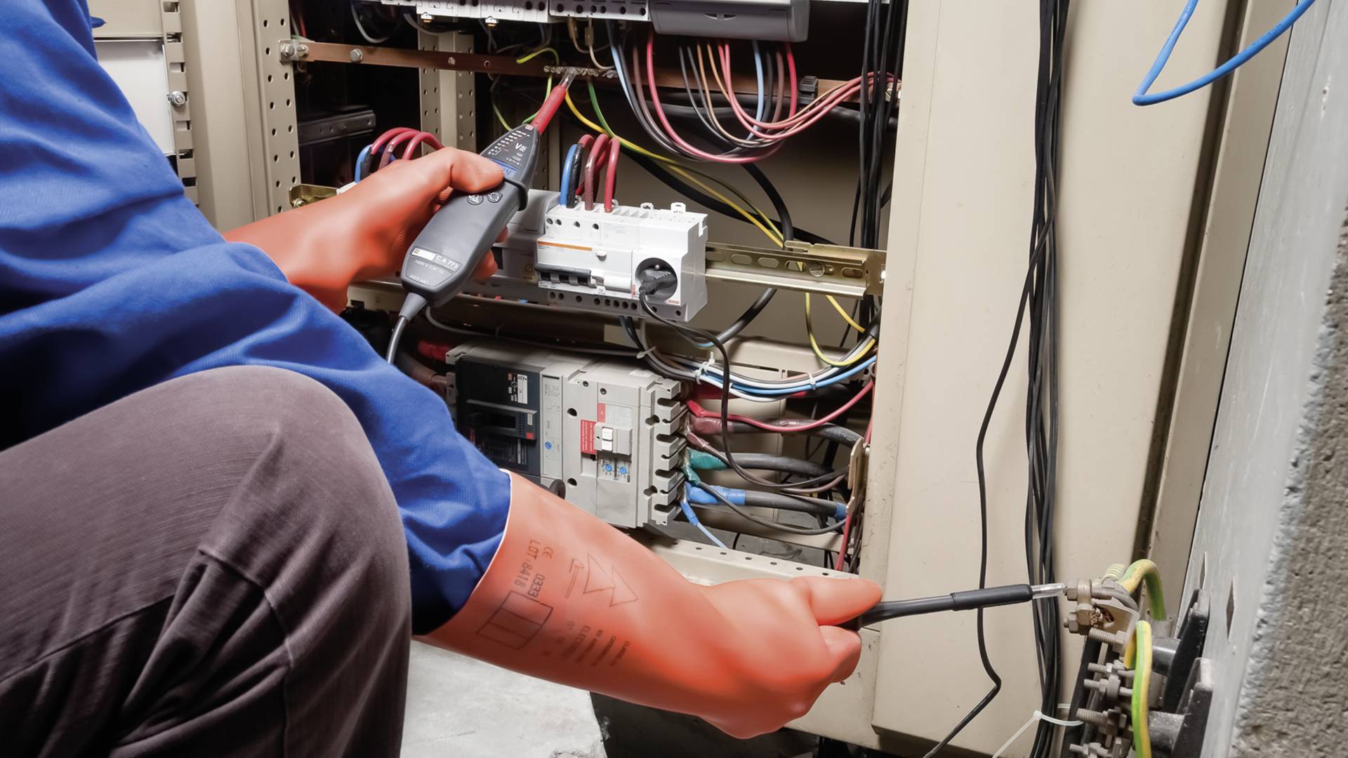 Elektryk w niebieskim ubraniu roboczym pracuje z narzędziem przy szafce sterowniczej pełnej kabli i elementów, wykonując naprawę lub czynności konserwacyjne.