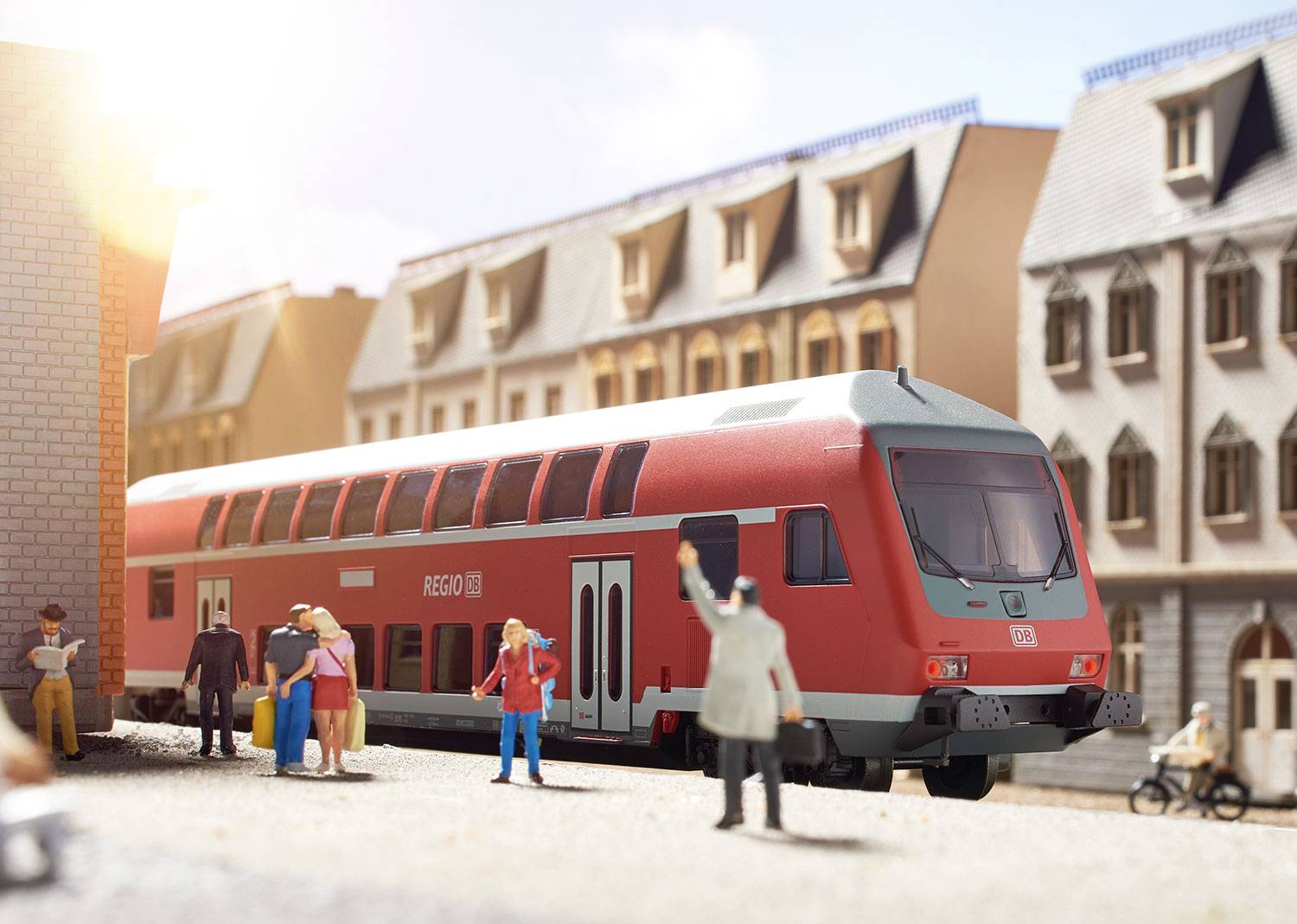 Czerwony pociąg regionalny stoi na stacji obok starych budynków. Ludzie z bagażami poruszają się po peronie. Jest słonecznie.