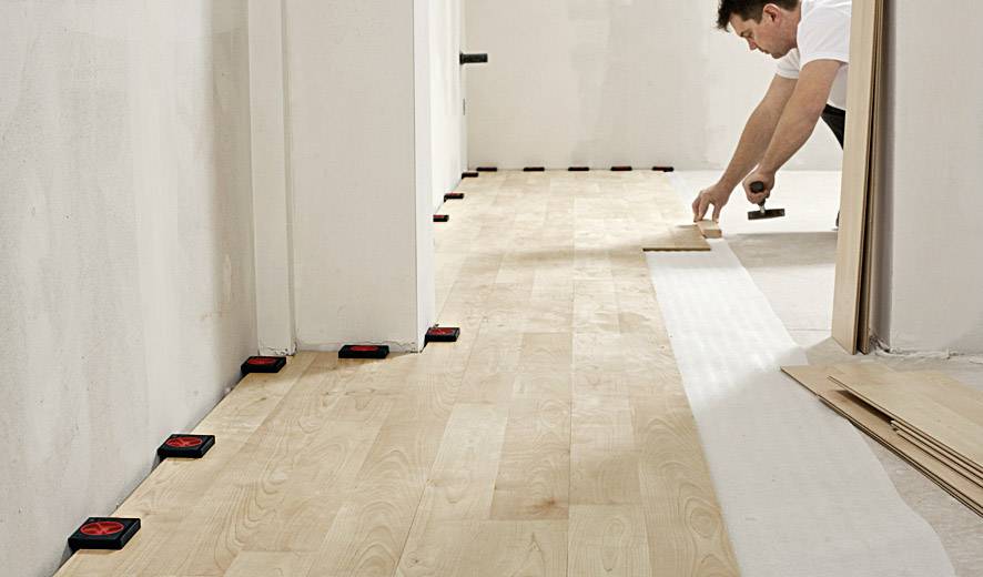 Osoba kładzie drewnianą podłogę w pomieszczeniu, używając młotka i dystansów do precyzyjnego rozmieszczenia paneli.