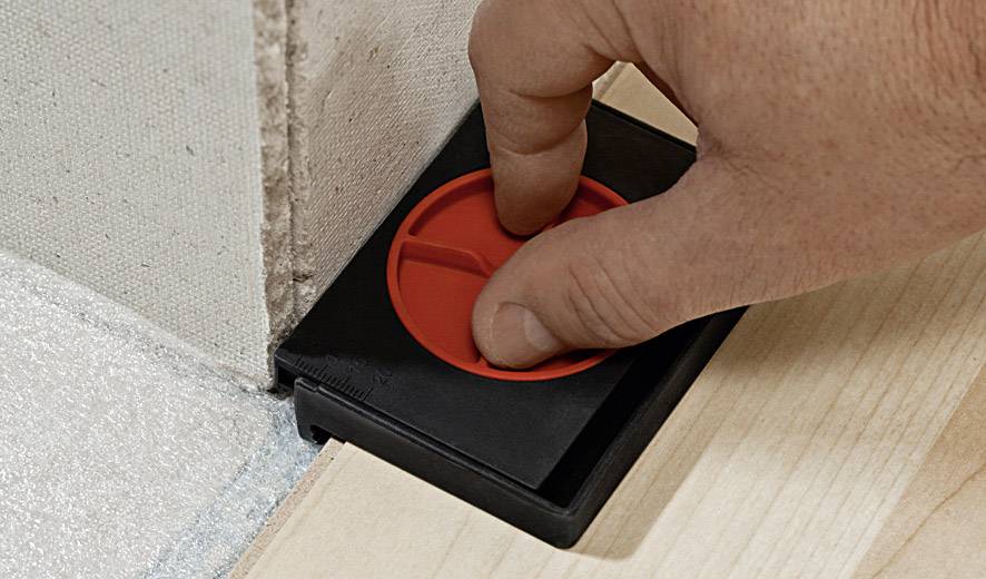 Dłoń naciska czerwony dystans między ścianą a drewnianą podłogą, aby zapewnić równomierną przerwę podczas instalacji.