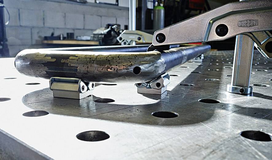 Metalowy rurociąg na stole warsztatowym z uchwytami, częściowo obrabiany. W tle widoczne są narzędzia i maszyny.