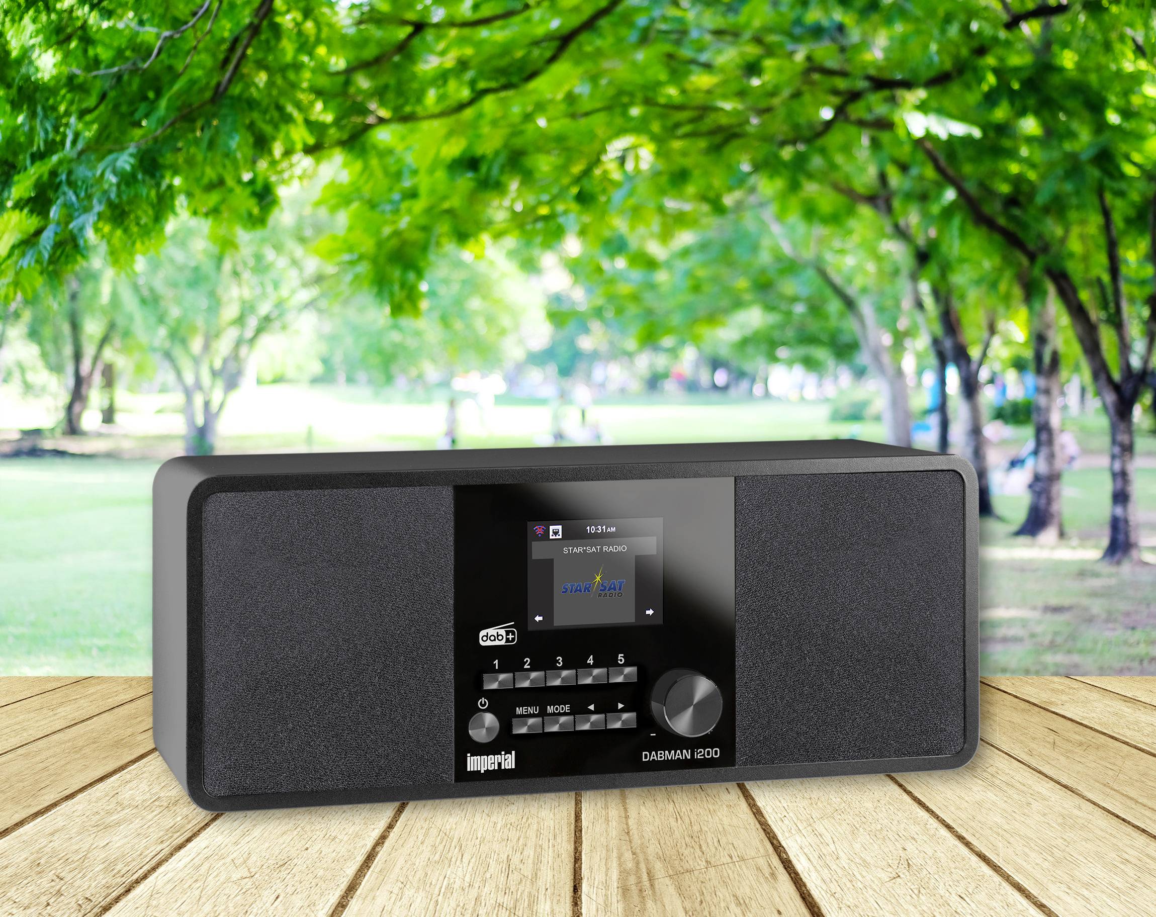 Radio stoi na drewnianym stole na tle zielonego krajobrazu parkowego. Na wyświetlaczu widać stację muzyczną.