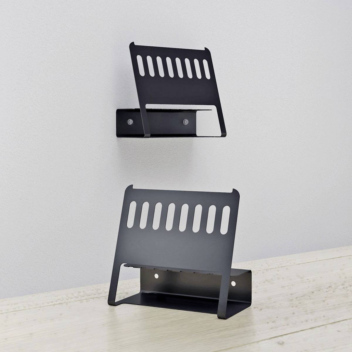 Czarne metalowe półki ścienne na jasnej ścianie, jedna wyższa, a druga niższa, każda z otworowanym wzorem i dwoma widocznymi otworami na wkręty.