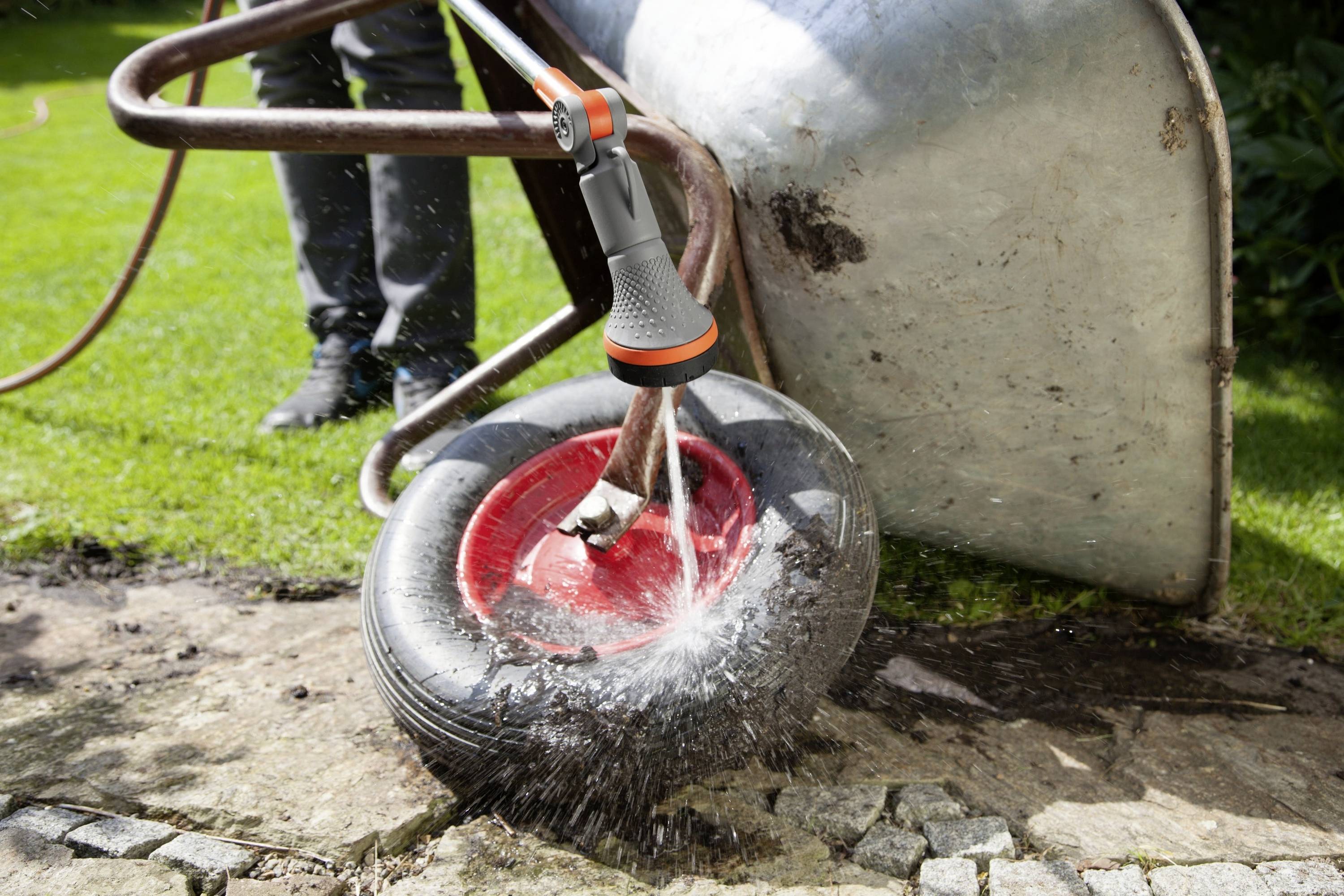 Osoba spryskuje koło taczki wodą z wyposażonego węża ogrodowego. Koło jest czerwono-czarne i mokre.