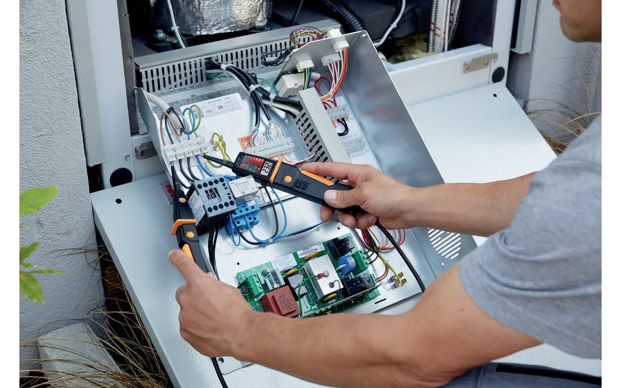 Osoba przeprowadza pomiary multimetrem na otwartej instalacji elektrycznej, prawdopodobnie w celu konserwacji lub naprawy.