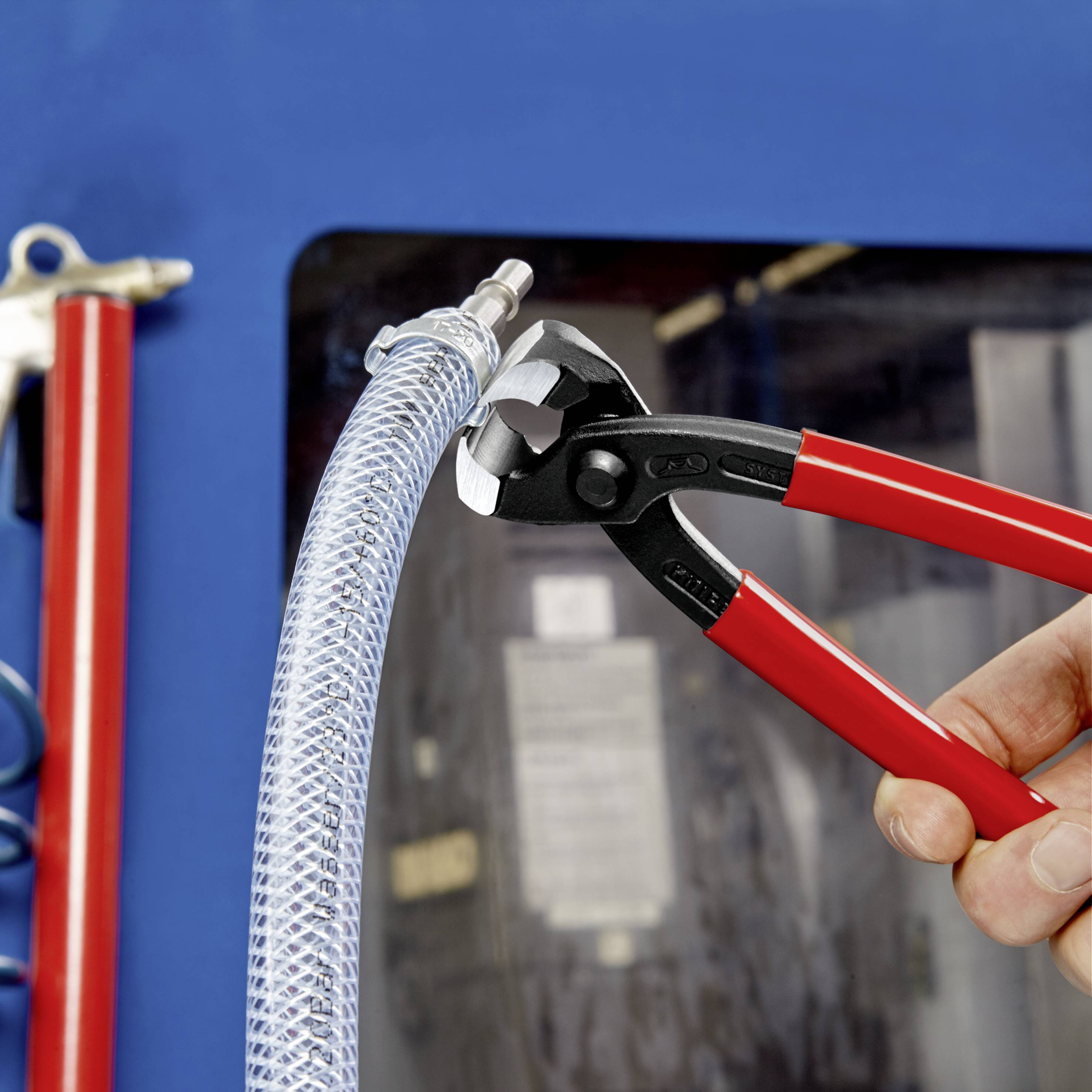 Ręka trzyma czerwone szczypce hydrauliczne, które mocno chwytają jasną, przezroczystą rurę. W tle niewyraźne maszyny.
