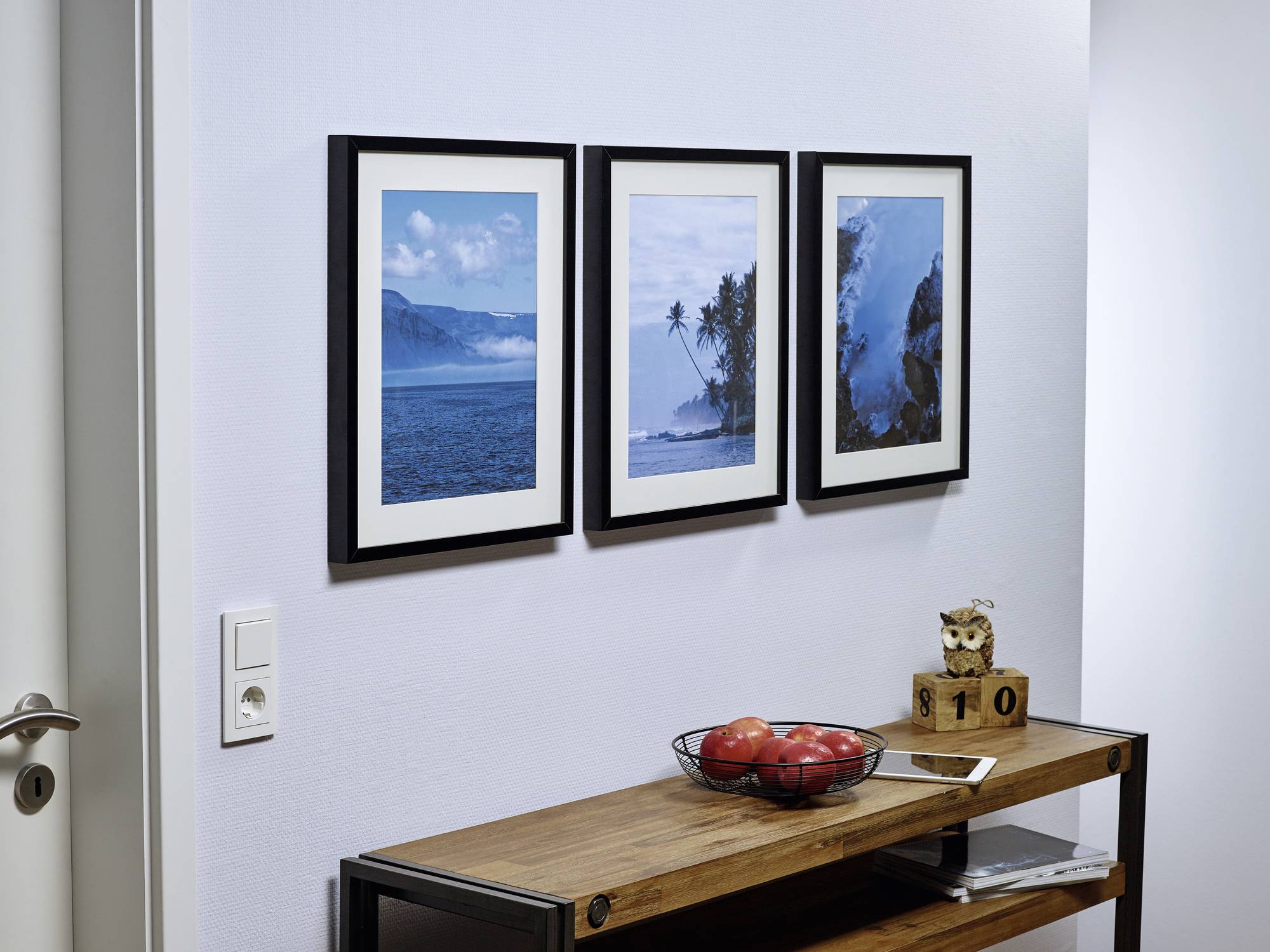 Trzy oprawione zdjęcia krajobrazowe na ścianie nad drewnianym stołem z koszem owoców i małym zegarem z figurką sowy.
