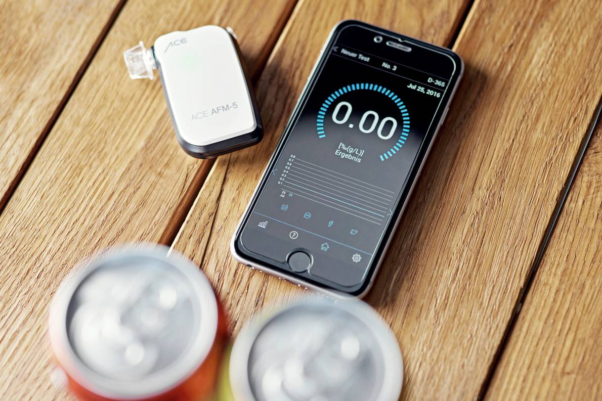 Smartfon na drewnianym stole wyświetla 0,00 promila na aplikacji do pomiaru poziomu alkoholu. Urządzenie obok. Na pierwszym planie nieostre puszki z napojami.