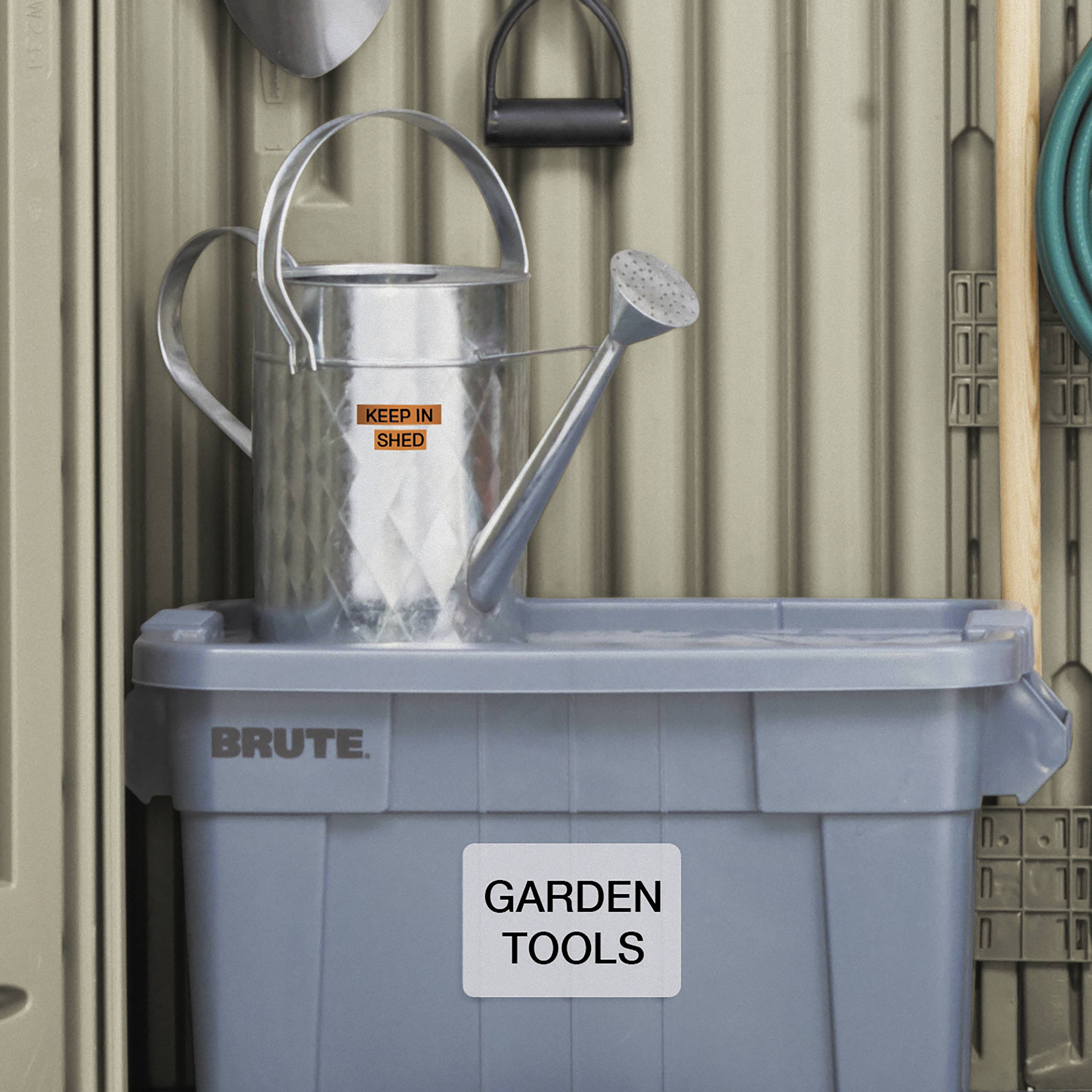 Szara plastikowa skrzynka z napisem 'Garden Tools' stoi przed ścianą, na której wisi konewka oraz haki z powieszonymi narzędziami ogrodowymi.