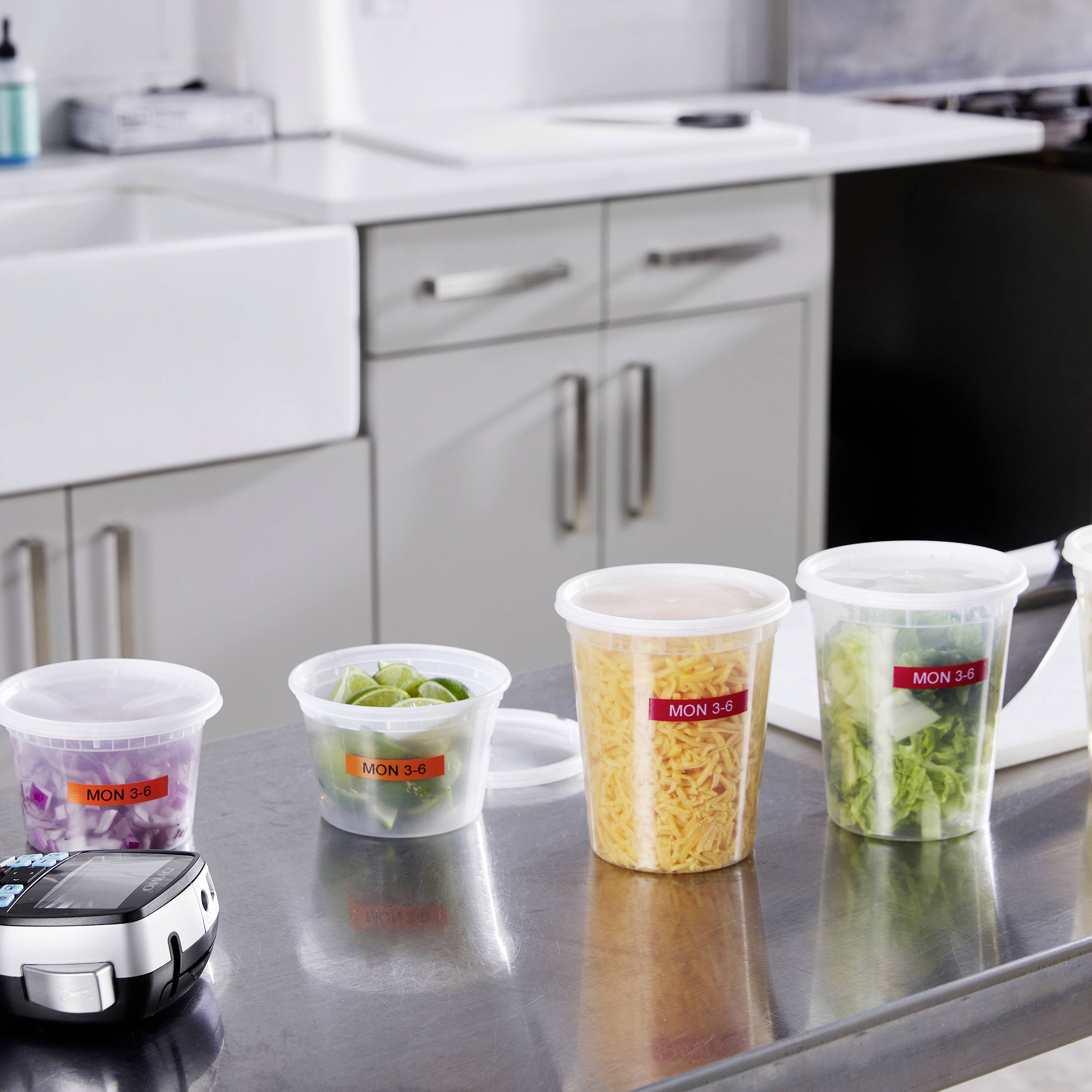 Kilka plastikowych pojemników z etykietą 'MON 3-6' ustawionych na stalowym blacie, wypełnionych sałatką i serem. W tle: szafki kuchenne.