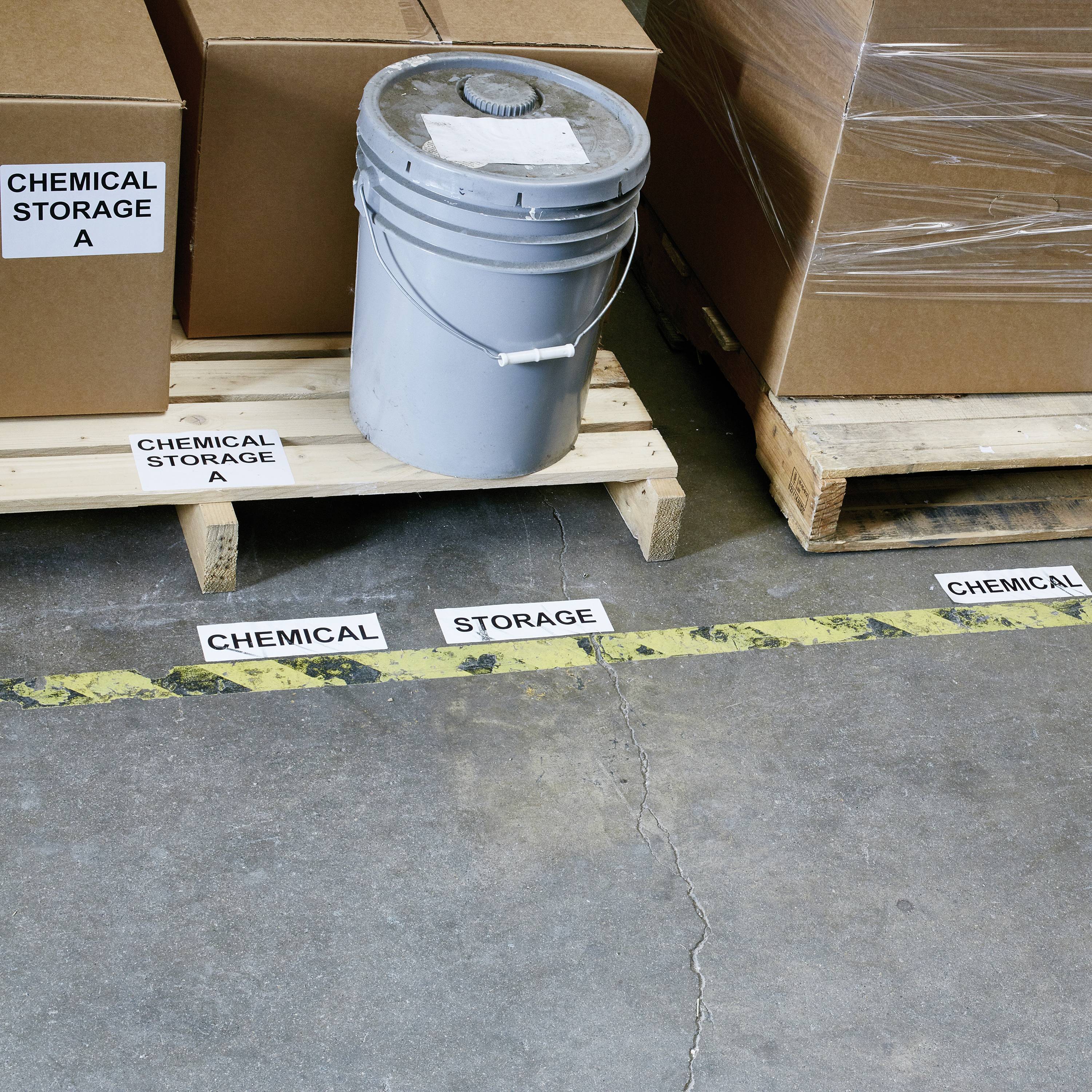 Magazyn z oznaczonymi tabliczkami na podłodze: 'MAGAZYN CHEMIKALIÓW', kartony i plastikowe wiadro na palecie.