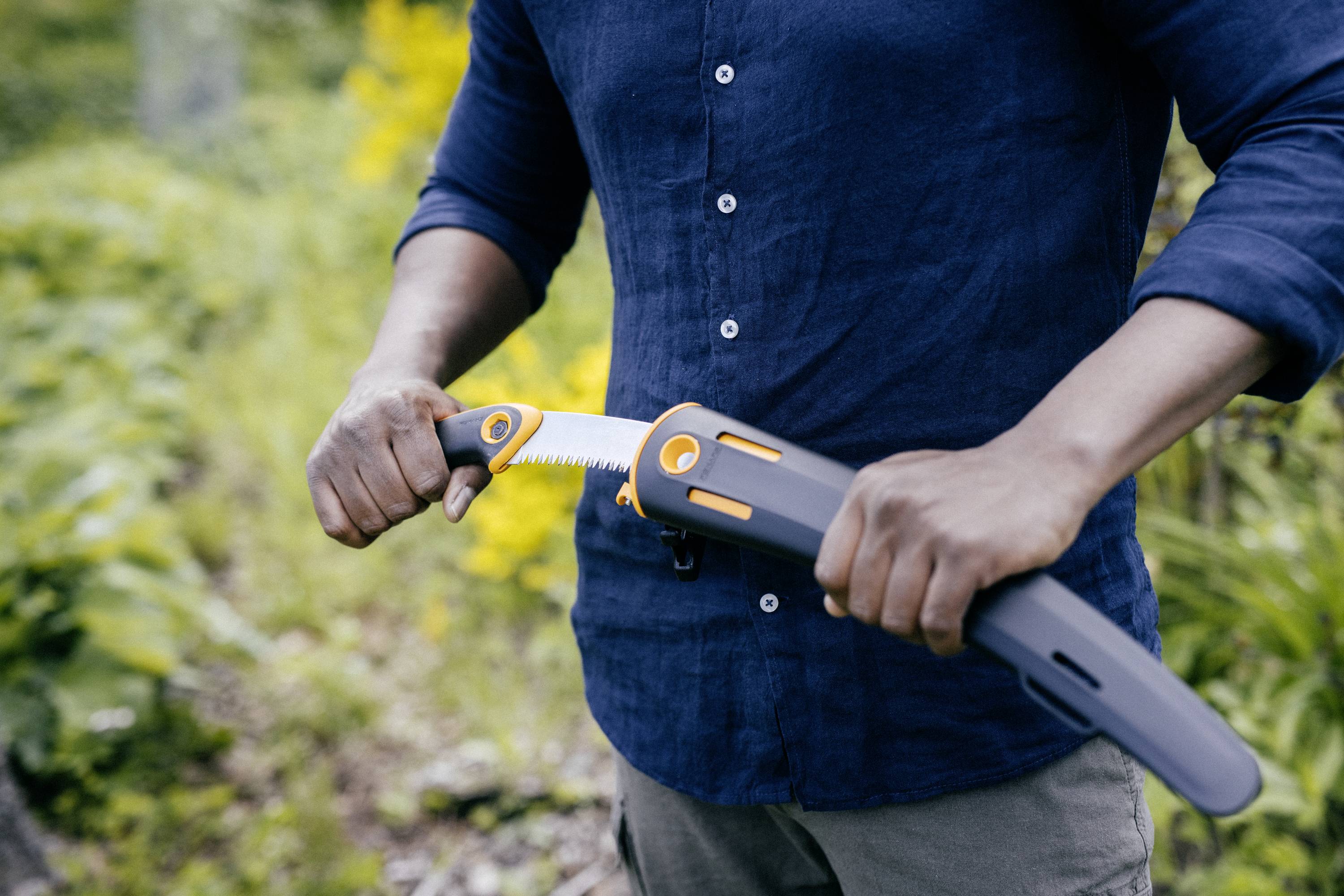 Osoba wyciąga piłę ręczną z uchwytu na porośniętej ścieżce. Osoba ma na sobie niebieską koszulę i trzyma piłę w bezpieczny sposób.