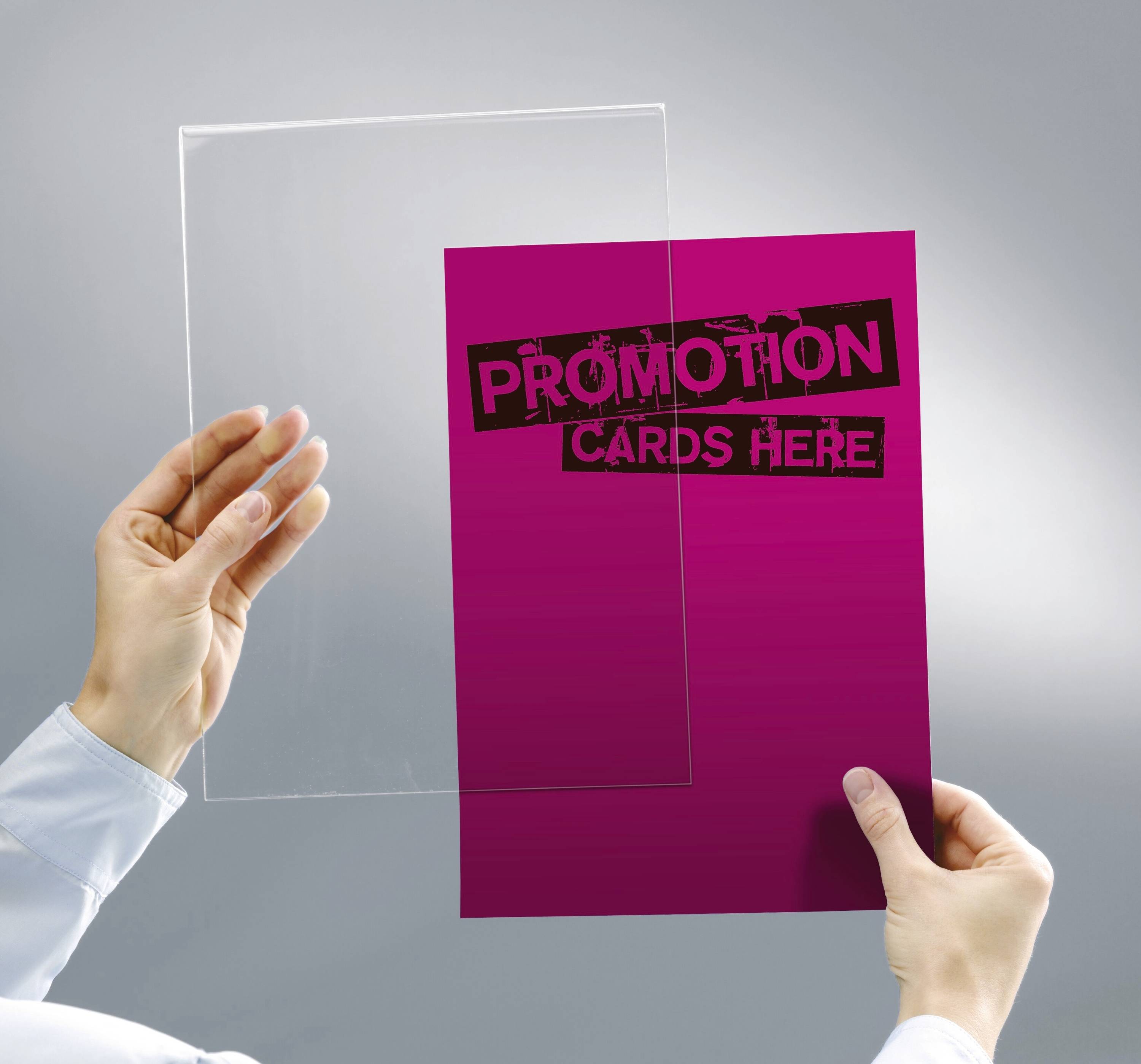 Ręce osoby trzymają przezroczystą folię i różową kartę z napisem "Karty promocyjne tutaj" na szarym tle.