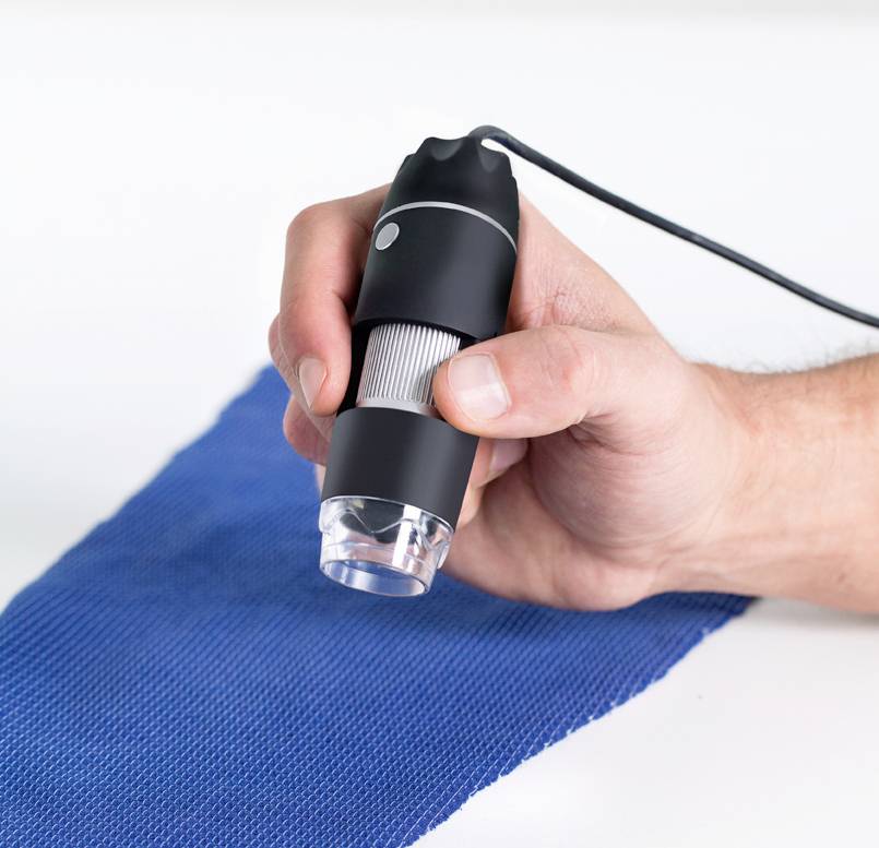 Jedna dłoń trzyma przenośny mikroskop nad niebieskim materiałem, aby zbadać jego strukturę.