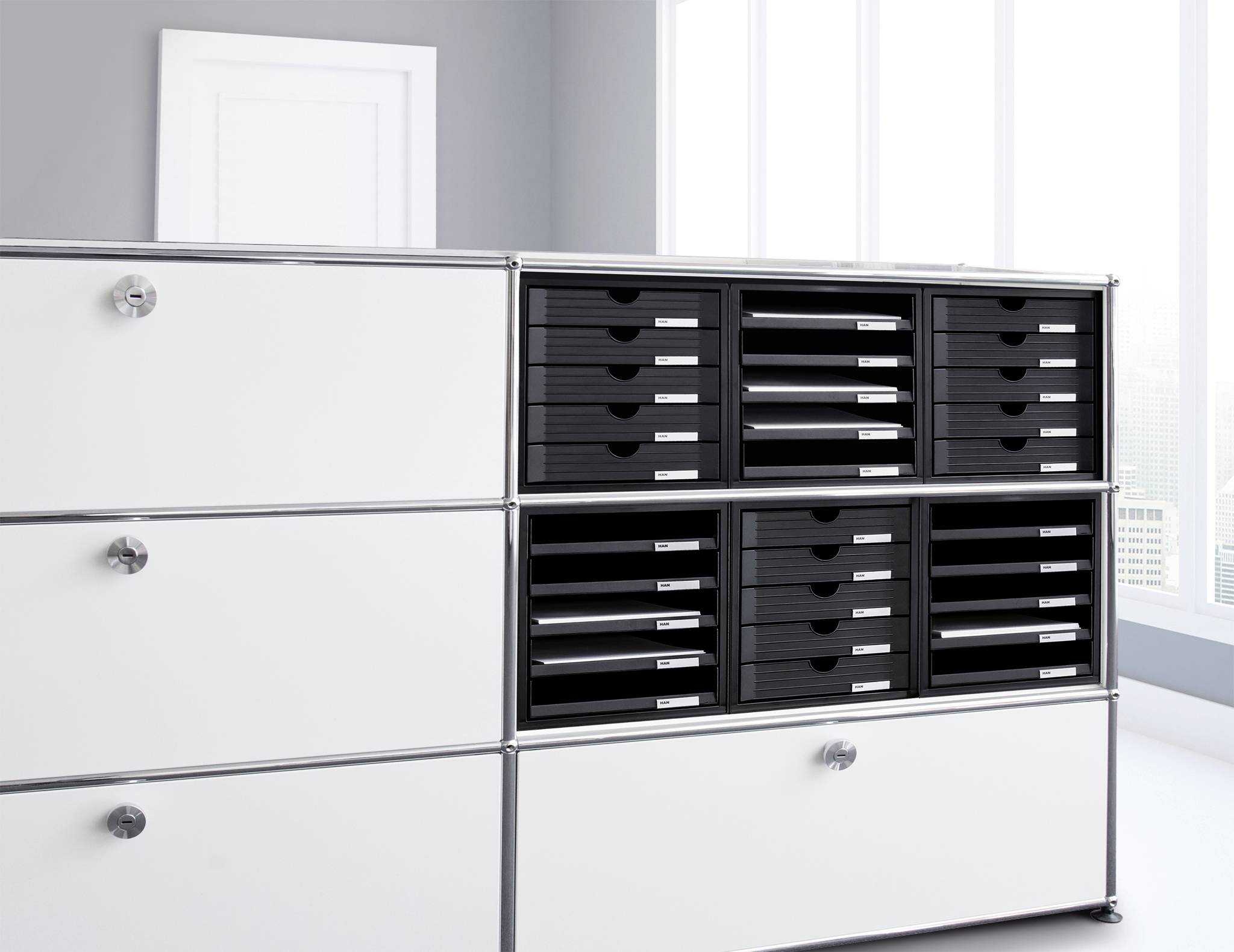 System regałowy z białymi drzwiami i otwartymi przegrodami, wypełniony czarnymi pojemnikami na dokumenty, w nowoczesnym, jasnym pomieszczeniu biurowym.