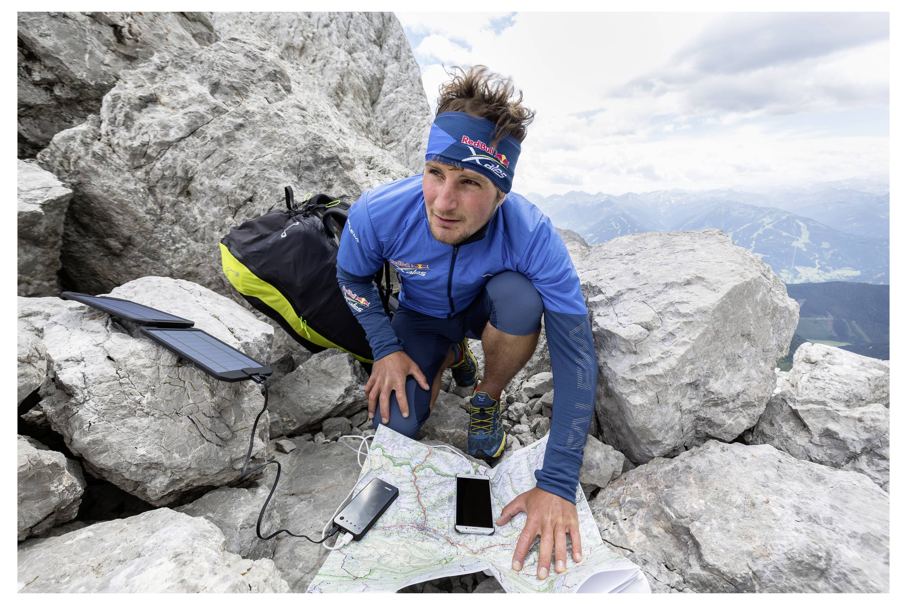 Osoba ubrana w niebieskie ubranie outdoorowe sprawdza mapę na skalistym terenie, w pobliżu znajdują się urządzenia elektroniczne i ładowarka słoneczna, a w tle widać góry.