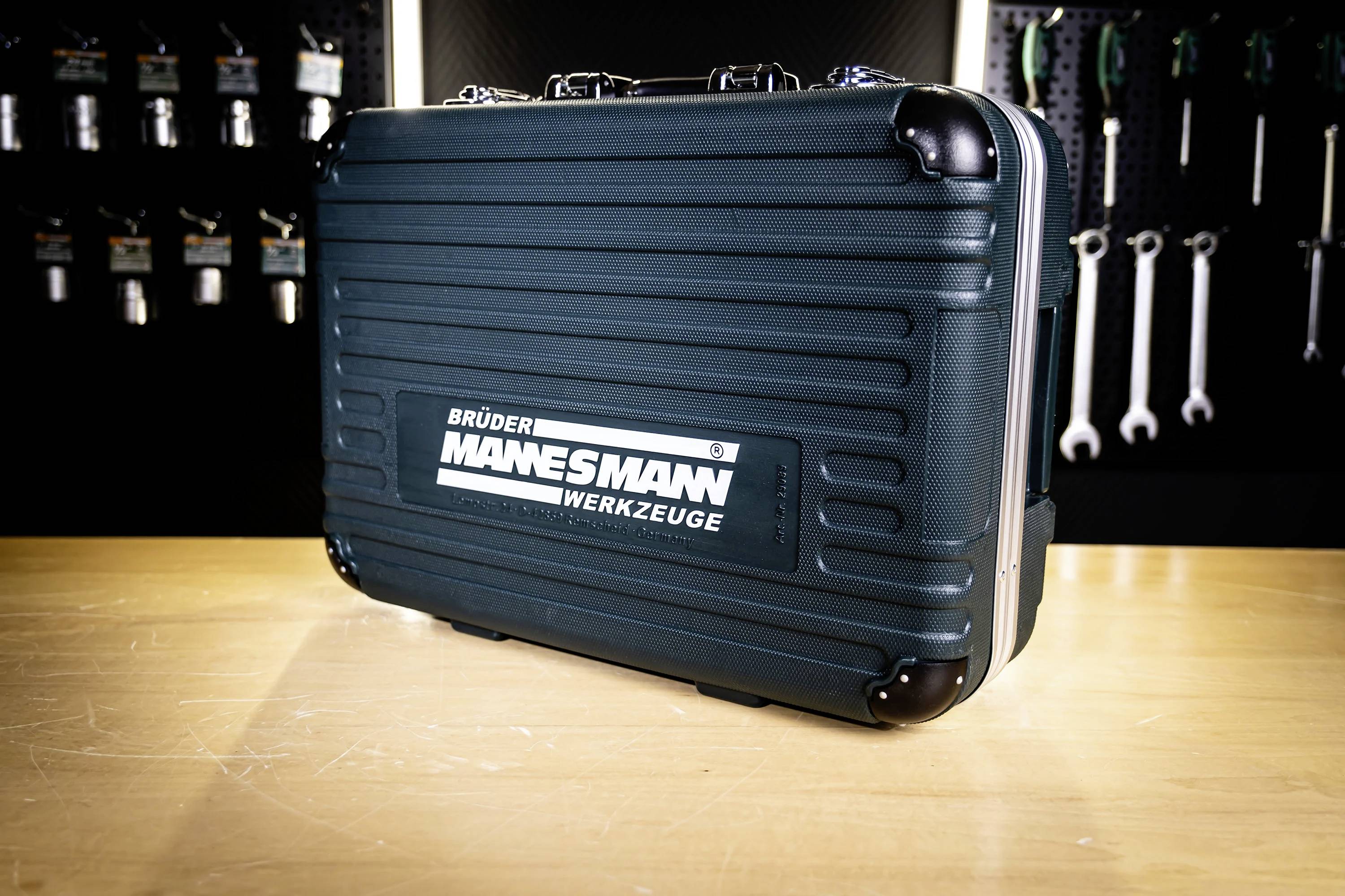 Czarna skrzynka narzędziowa z etykietą 'Brüder Mannesmann Werkzeuge' na drewnianym stole. W tle widoczne są wiszące narzędzia na ścianie.