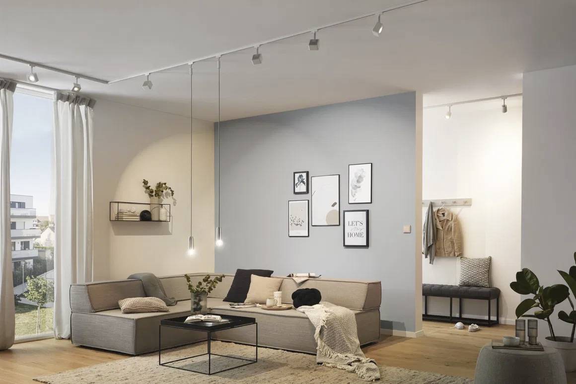 Jasny salon z szarą sofą, dekoracyjnymi ramkami na ścianach i nowoczesnymi lampami. Na zewnątrz balkon z widokiem na ogród.