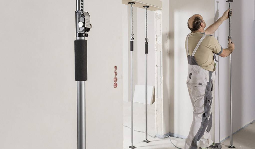 Robotnik instaluje podporę przy ścianie, w tle widać kolejne podpory. Jest ubrany w odzież ochronną i klęczy.