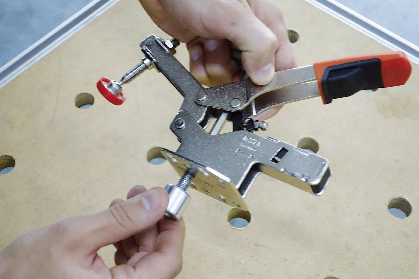 Jedna ręka trzyma drewnianą szczypce montażowe z pomarańczową rękojeścią. Druga ręka przygotowuje srebrną śrubę do zamocowania.