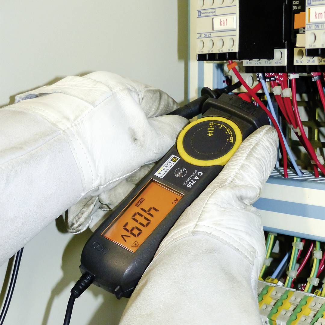 Technik w białych rękawicach ochronnych mierzy napięcie w elektrycznej szafie rozdzielczej za pomocą cyfrowego multimetru, który wskazuje 408 woltów.