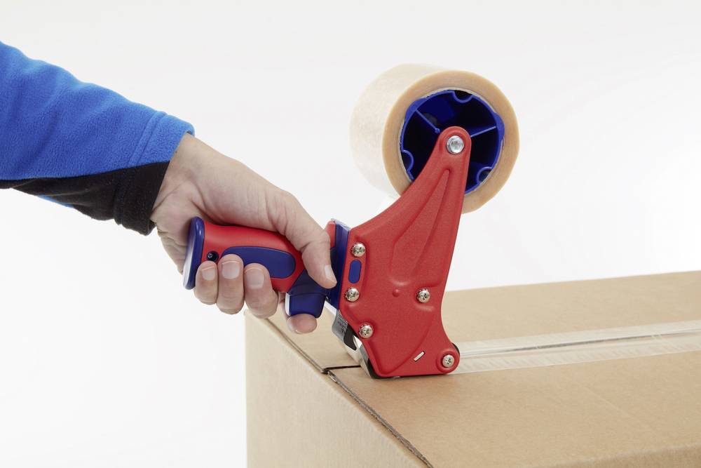 Osoba zamyka karton za pomocą ręcznego dyspensera do taśmy klejącej w kolorze czerwonym, który jest umieszczony na rogu kartonu.