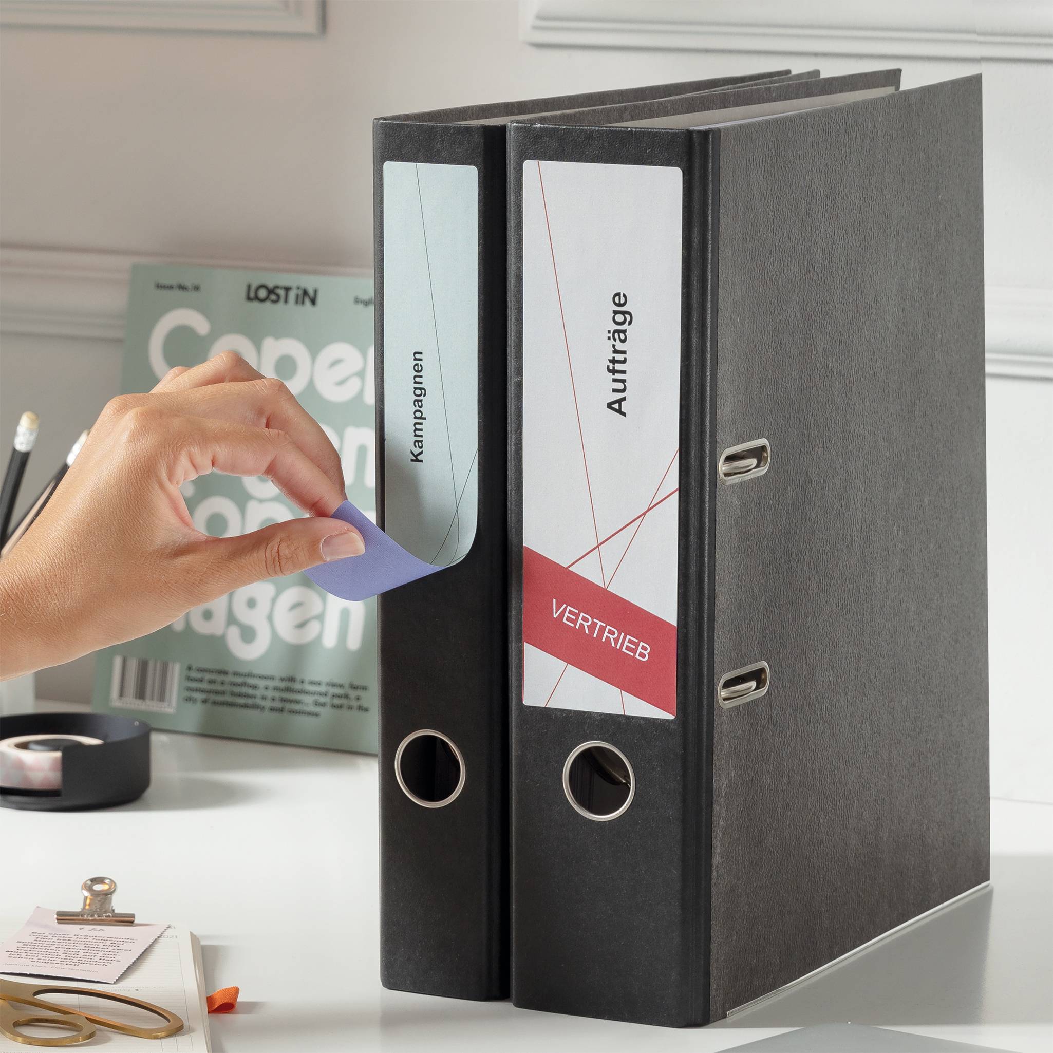 Ręka przykleja niebieski arkusz na czarną teczkę z napisem 'Kampanie', obok znajduje się segregator z napisem 'Zlecenia'. W tle widać czasopismo i przybory biurowe.