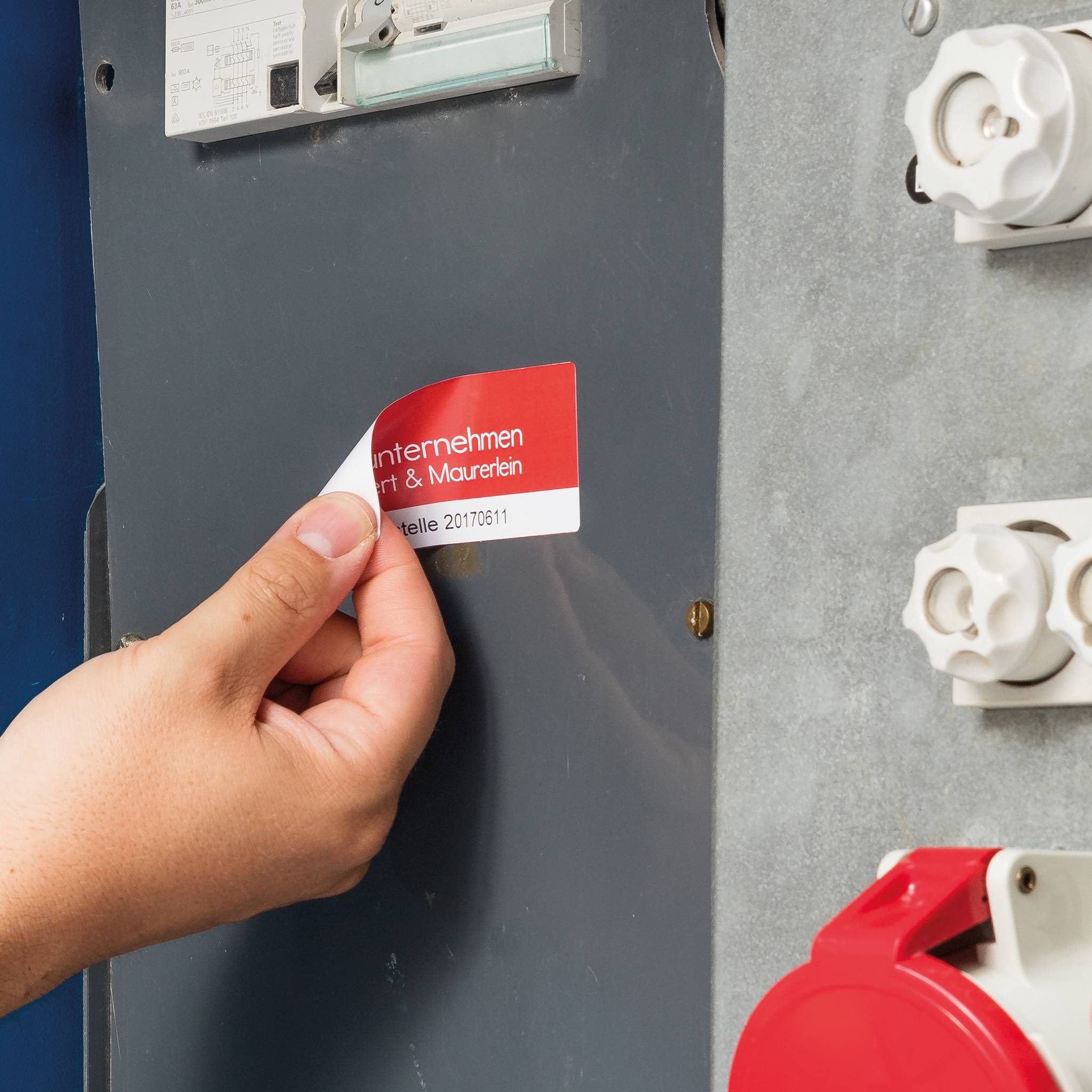 Ręka zdejmuje czerwoną naklejkę z szarej szafki elektrycznej. Na naklejce widnieje napis 'przedsiębiorstwo | Malarz & Murarz'.