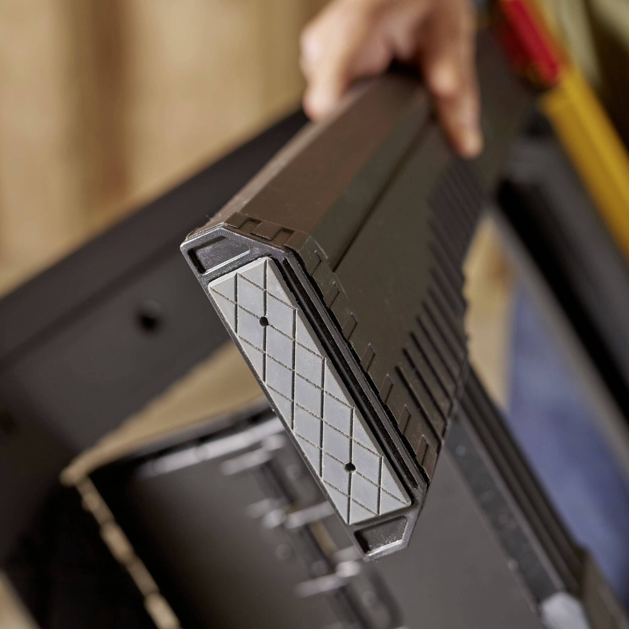 Dłoń trzyma czarny plastikowy element o karbowanej krawędzi. W tle widać niewyraźne materiały budowlane.