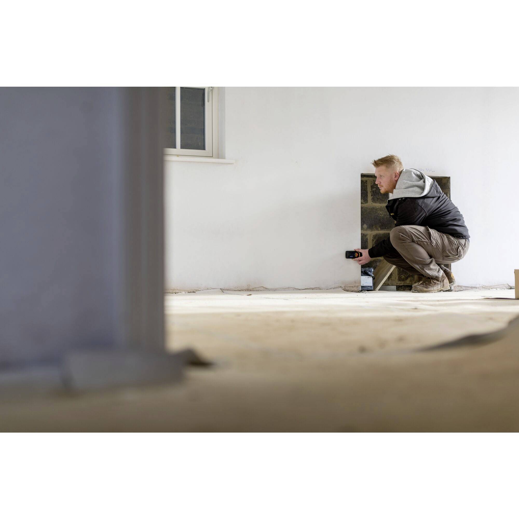 Mężczyzna w pustym pomieszczeniu, trzymający poziomicę przy ścianie, wyraźnie zajęty remontem. Skupiony na dokładnym wyrównaniu.