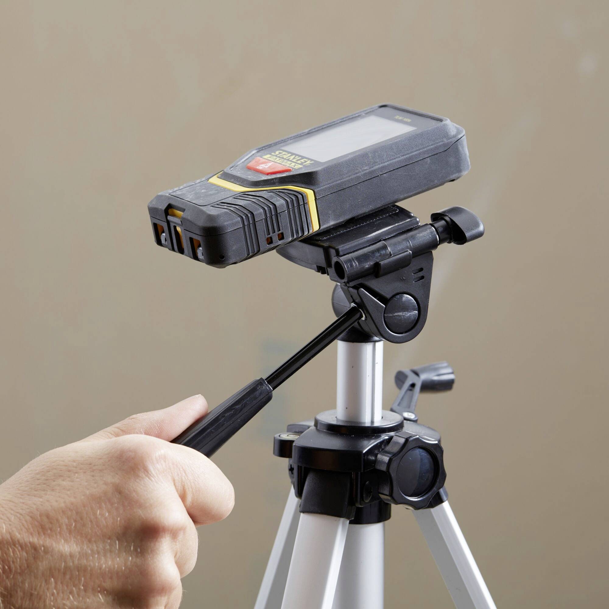 Jedna ręka trzyma cyfrowy laserowy miernik odległości na statywie, gotowy do pomiaru we wnętrzu.