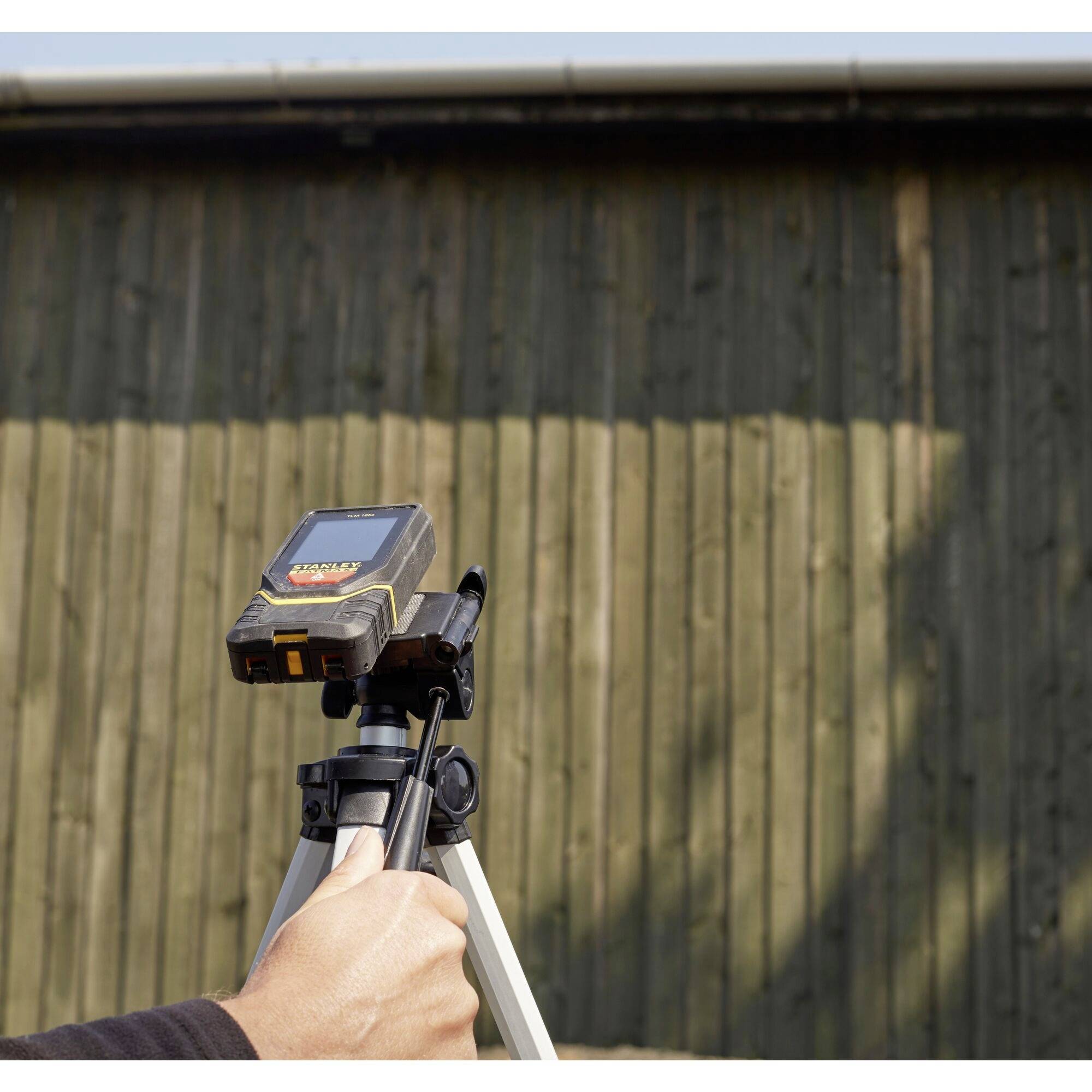 Dokonywany jest pomiar odległości laserem. Urządzenie zamocowane na statywie wyświetla wartości pomiarowe, podczas gdy mężczyzna trzyma je przed drewnianą ścianą.