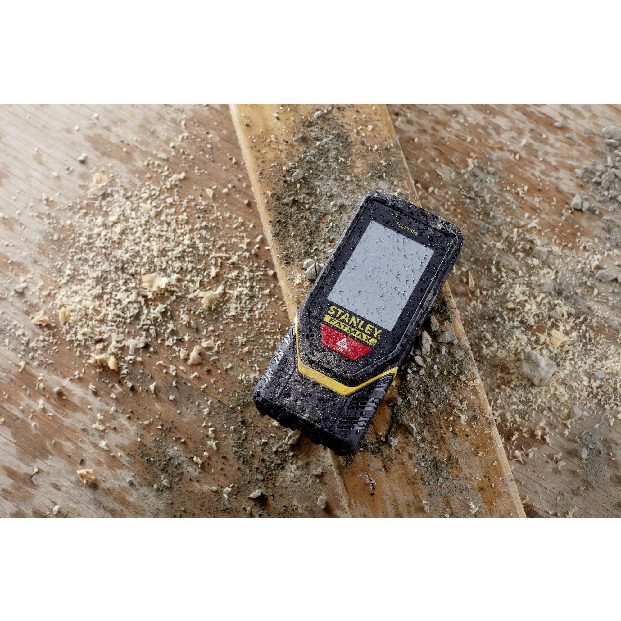 Laserowy miernik odległości leży na brudnej, drewnianej desce posypanej troczynami. Urządzenie ma oznaczenie marki 'Stanley'.