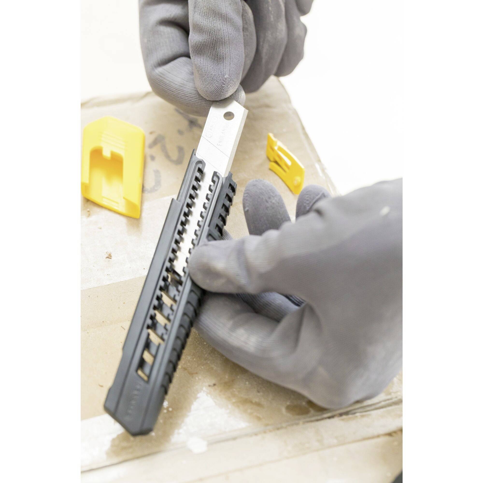 Osoba ubrana w szare rękawiczki trzyma nóż introligatorski nad kartonem. Obok leży kilka żółtych plastikowych osłonek.