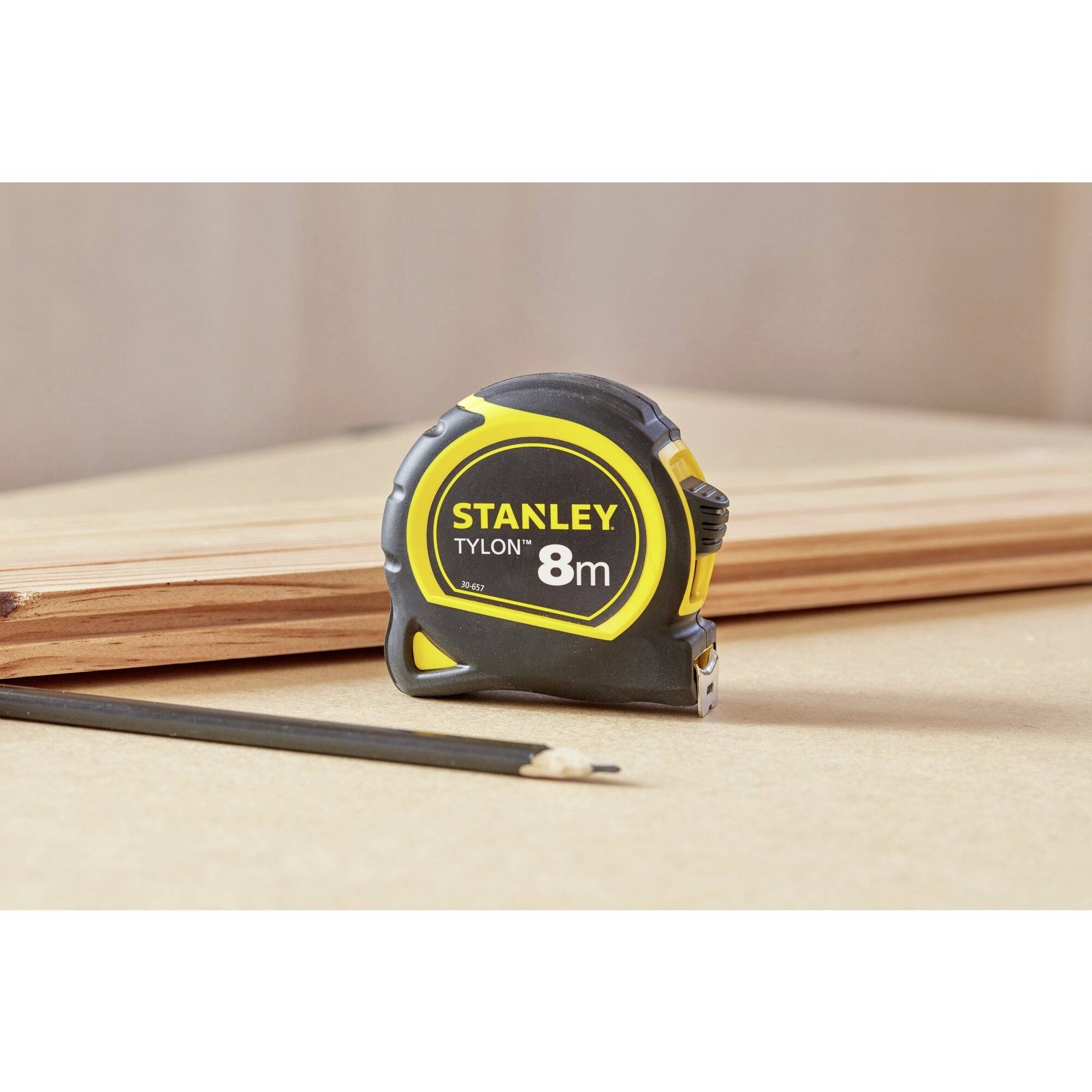Miara zwijana z napisem 'STANLEY TYLON 8m' leży na drewnianej desce obok ostro naostrzonego ołówka.