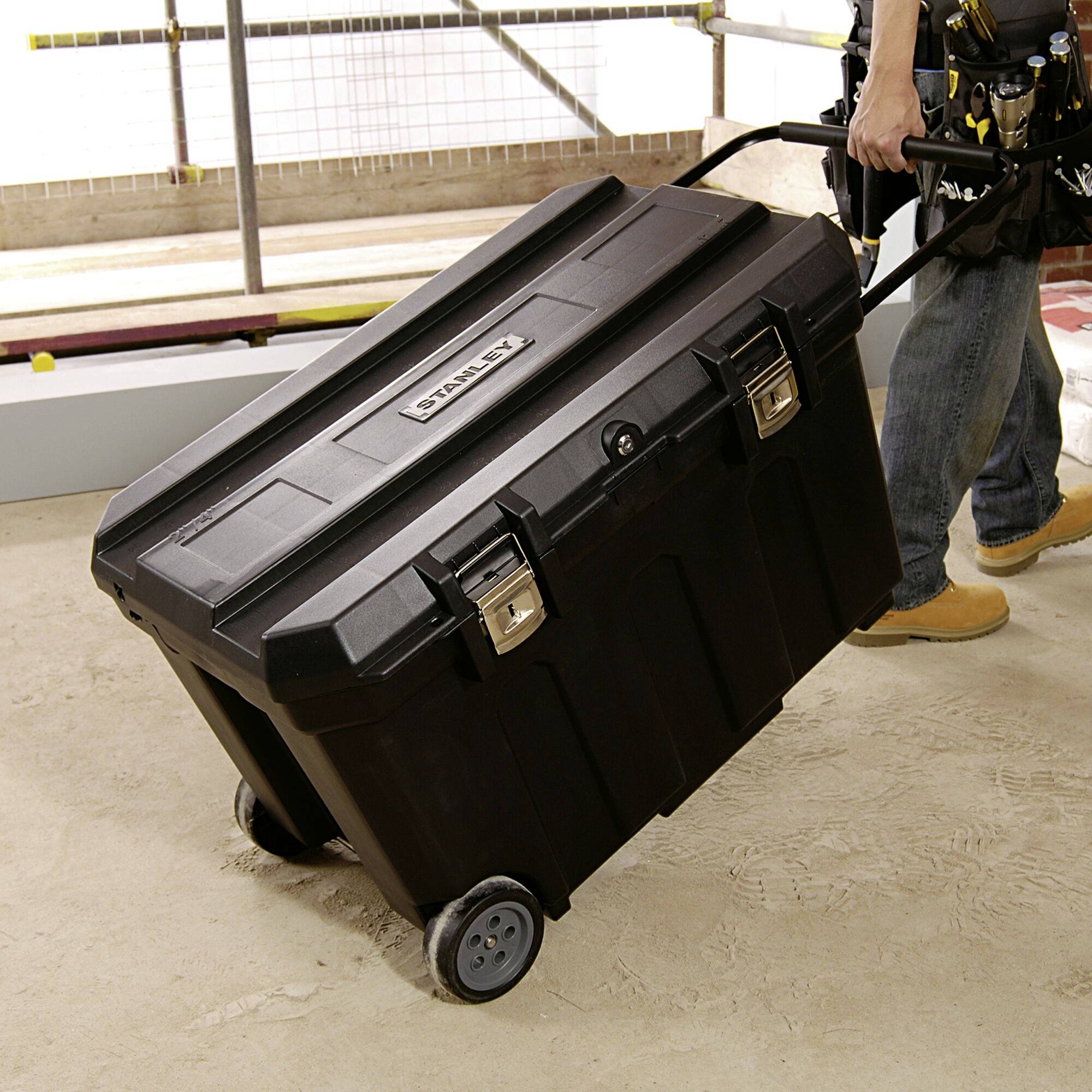 Osoba ciągnie dużą, czarną skrzynkę narzędziową na kółkach przez teren budowy. Skrzynka posiada srebrne zatrzaski.