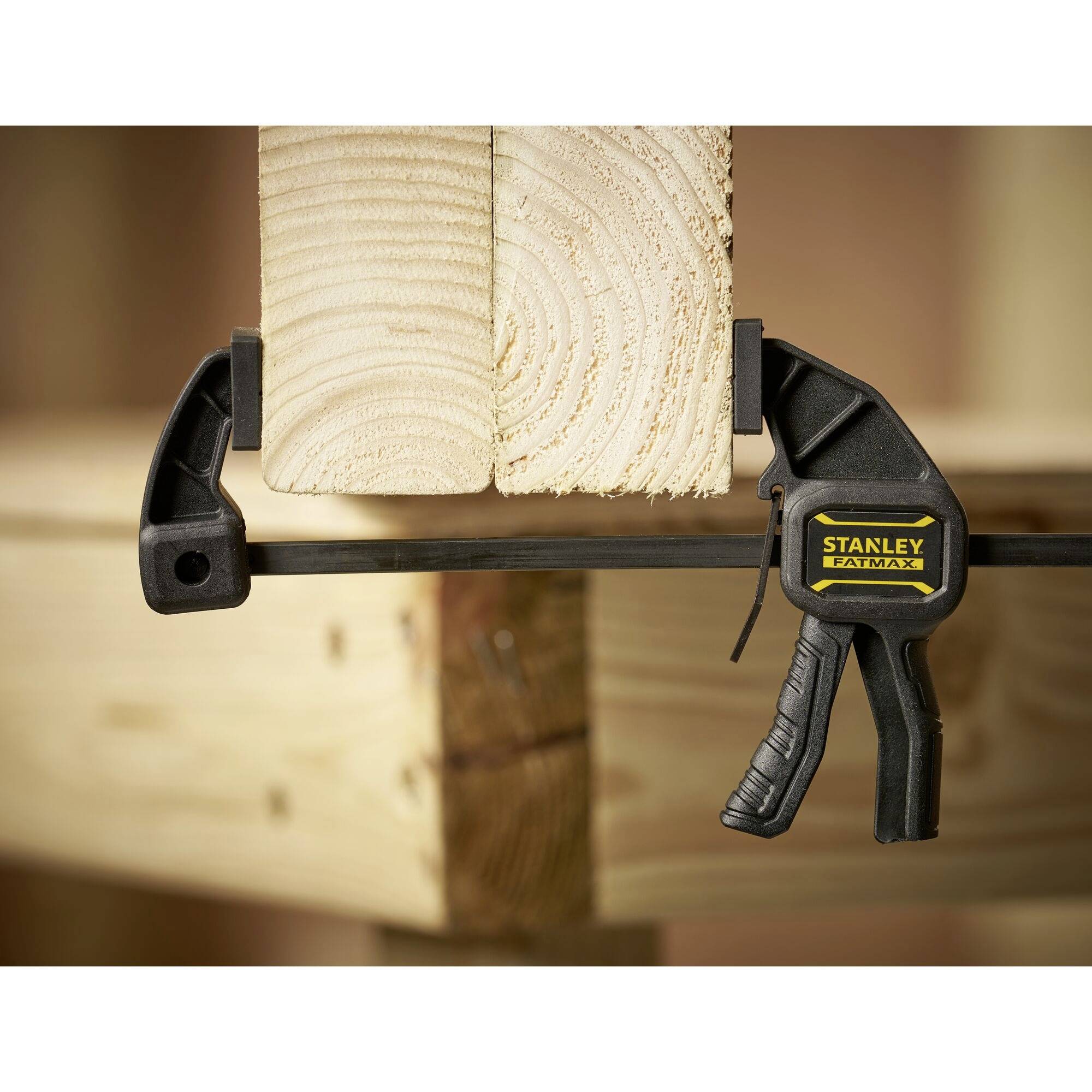 Scena przedstawiająca zacisk śrubowy mocujący dwie drewniane deski na stole. Zacisk posiada logo 'STANLEY Fatmax'.
