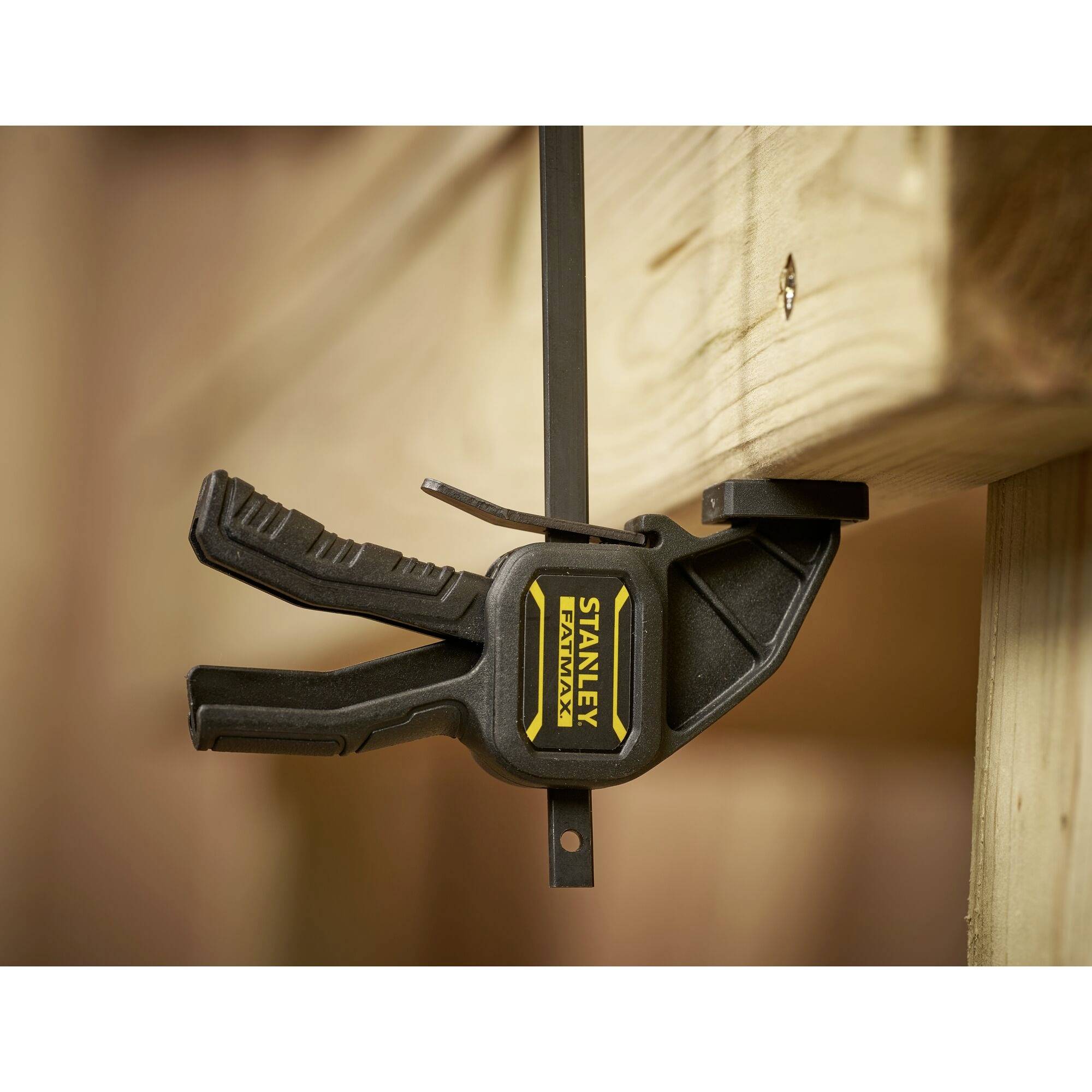 Czarna plastikowa szczęka zaciskowa mocuje kawałek drewna do stołu. Na szczęce widnieje napis 'STANLEY FATMAX'.