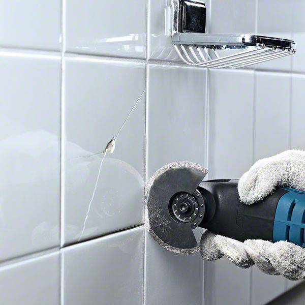 Jedna ręka trzyma narzędzie i ucina uszkodzoną płytkę w łazience, przygotowując się do naprawy pęknięcia.