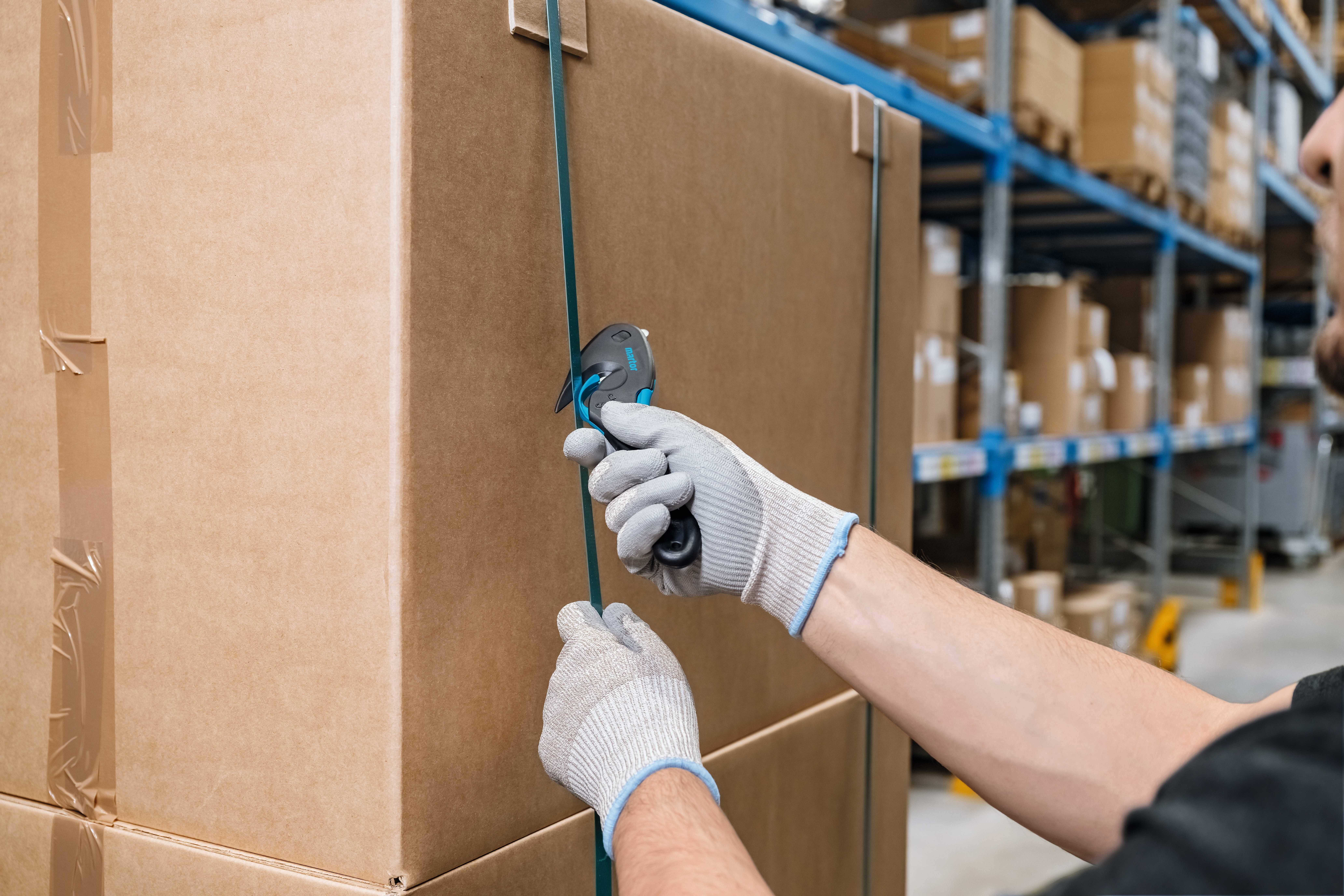 Osoba w rękawicach przecina plastikową taśmę owijającą duży karton w magazynie. W tle widoczne są regały z kartonami.