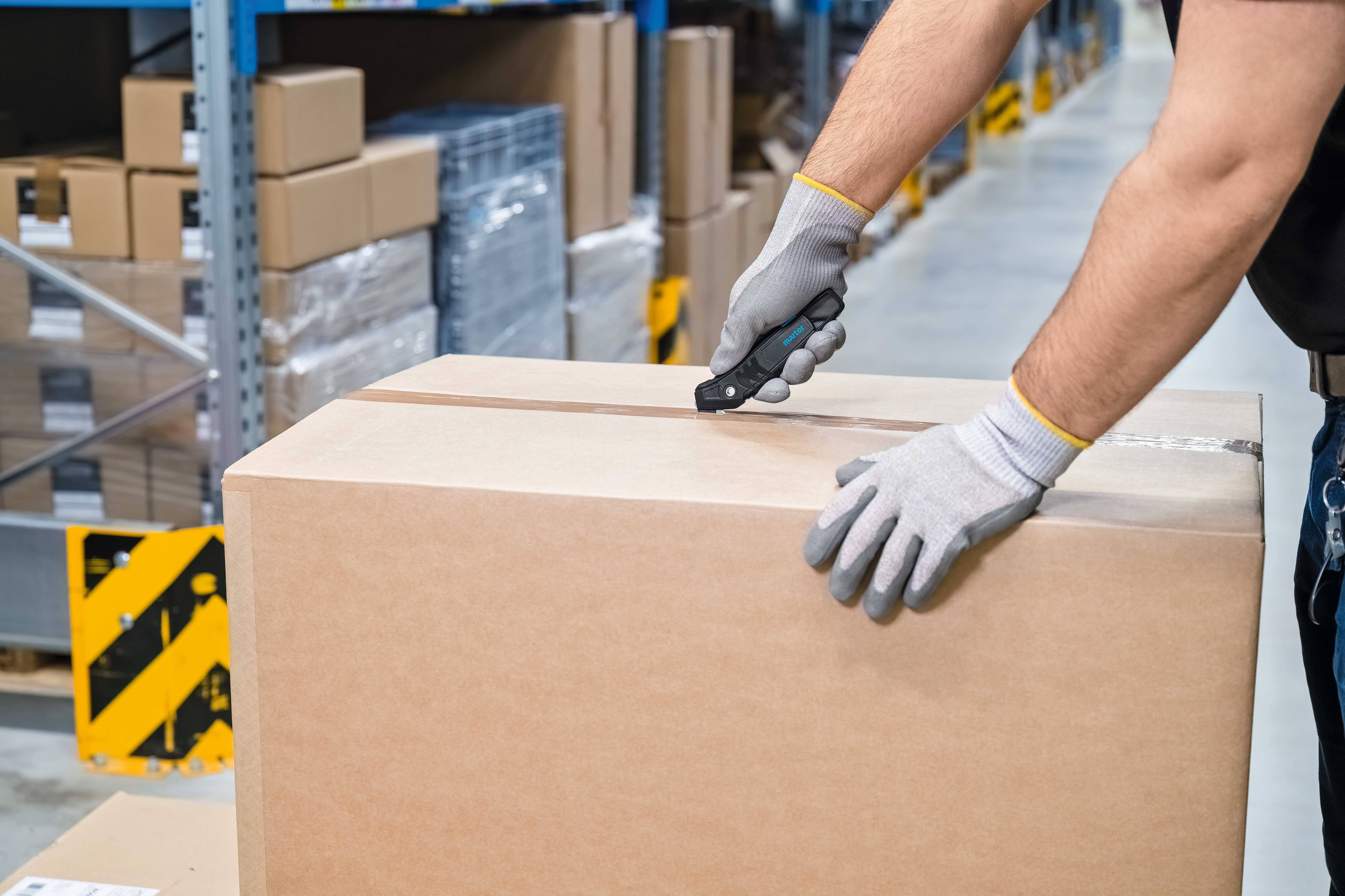 Osoba w rękawicach roboczych otwiera nożem duży karton w magazynie z regałami pełnymi paczek.