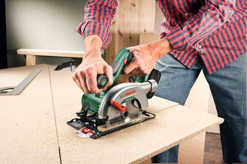 Osoba piłuje drewno zieloną ręczną piłą tarczową na stole w warsztacie. Otoczenie pokryte jest trocинами i płytami drewnianymi.