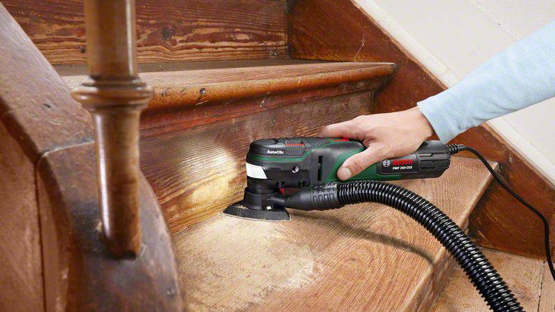Jedna osoba szlifuje drewniane schody wielofunkcyjnym narzędziem. Urządzenie jest podłączone do odkurzacza, aby usuwać pył.