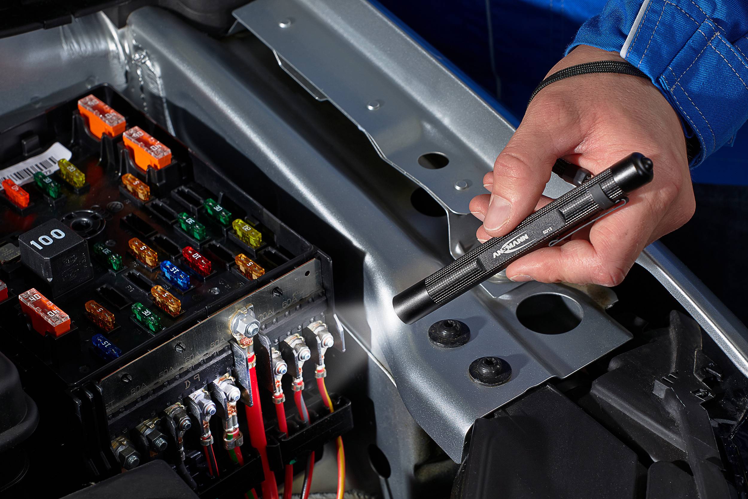 'Jedna ręka trzyma latarkę nad otwartą skrzynką bezpieczników w samochodzie. Widoczne są różnokolorowe bezpieczniki.'