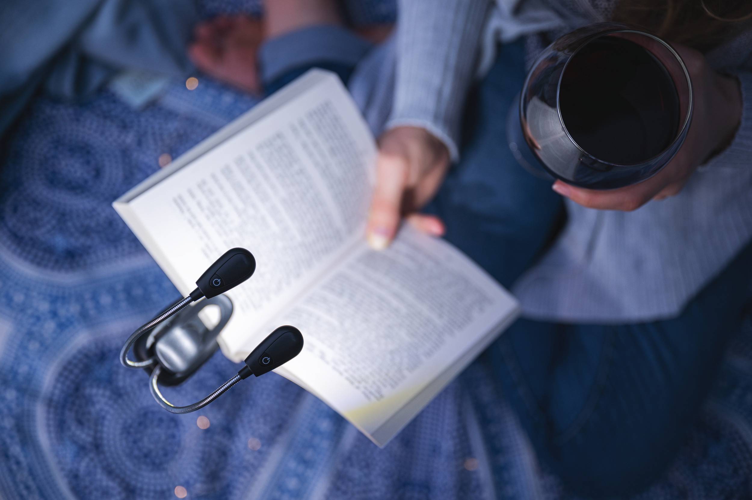 Osoba czyta książkę na ozdobnej tkaninie, oświetlona lampką do czytania, trzymając w drugiej ręce kieliszek czerwonego wina.