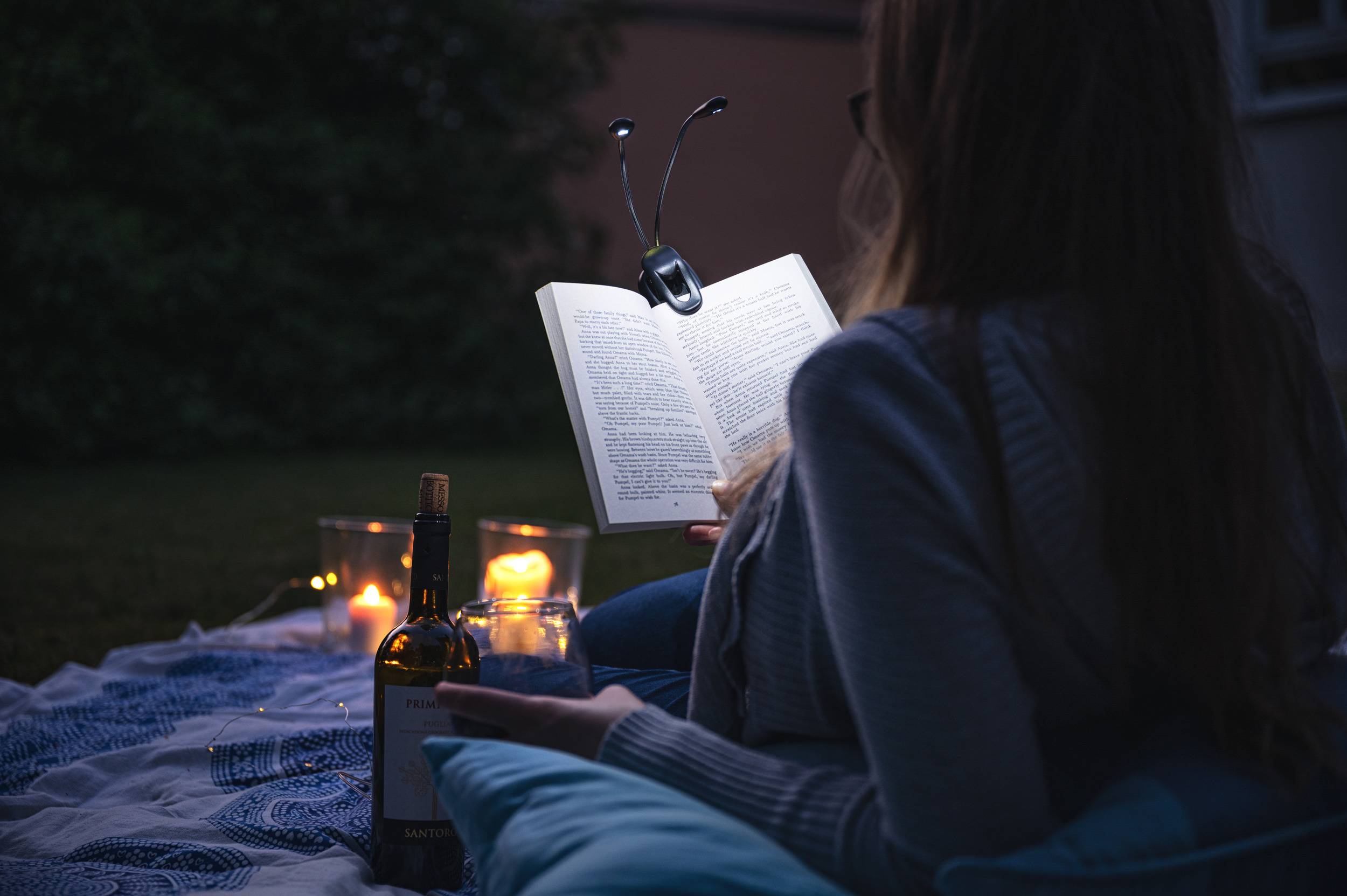 Osoba czyta książkę przy świecach na dworze. Obok niej stoją butelka wina i kieliszki. Panuje zrelaksowana atmosfera.