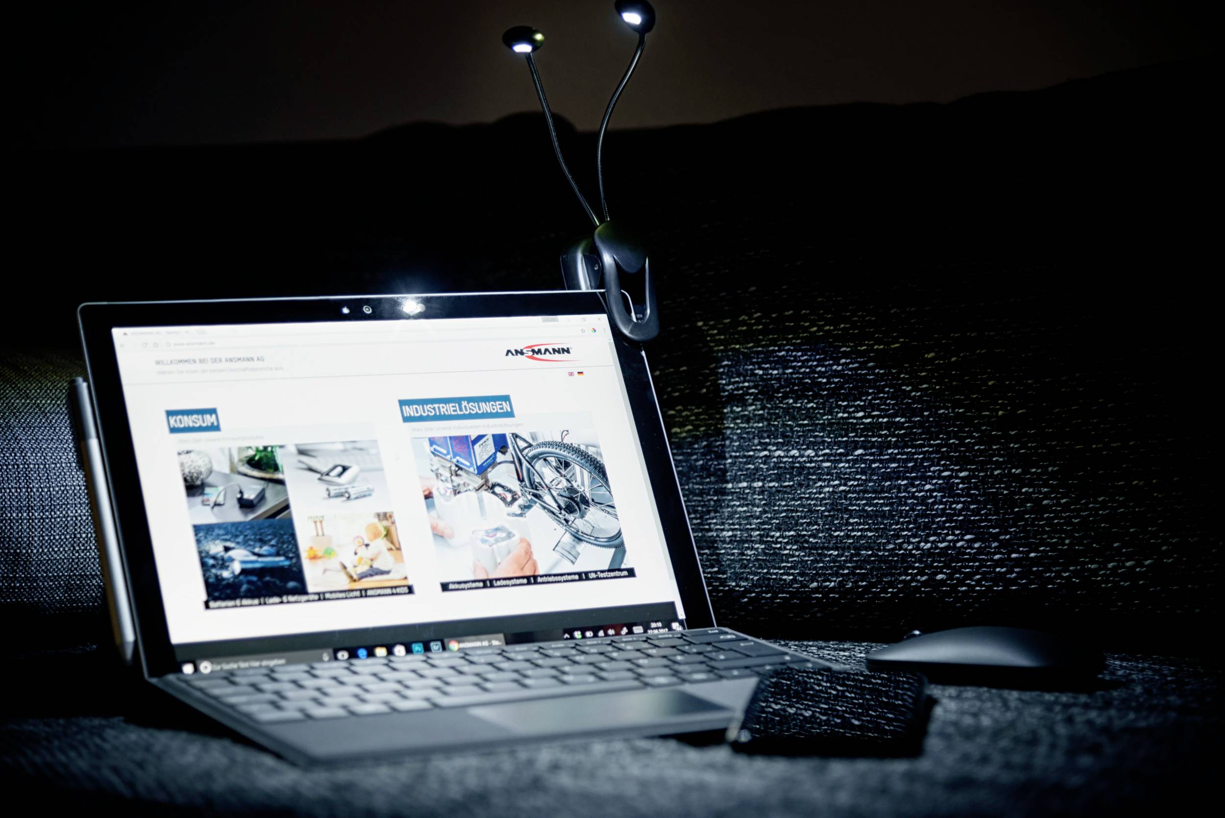 Laptop na kanapie wyświetla stronę internetową z różnymi kategoriami. Lampa świeci na klawiaturę, a obok niej leży mysz.