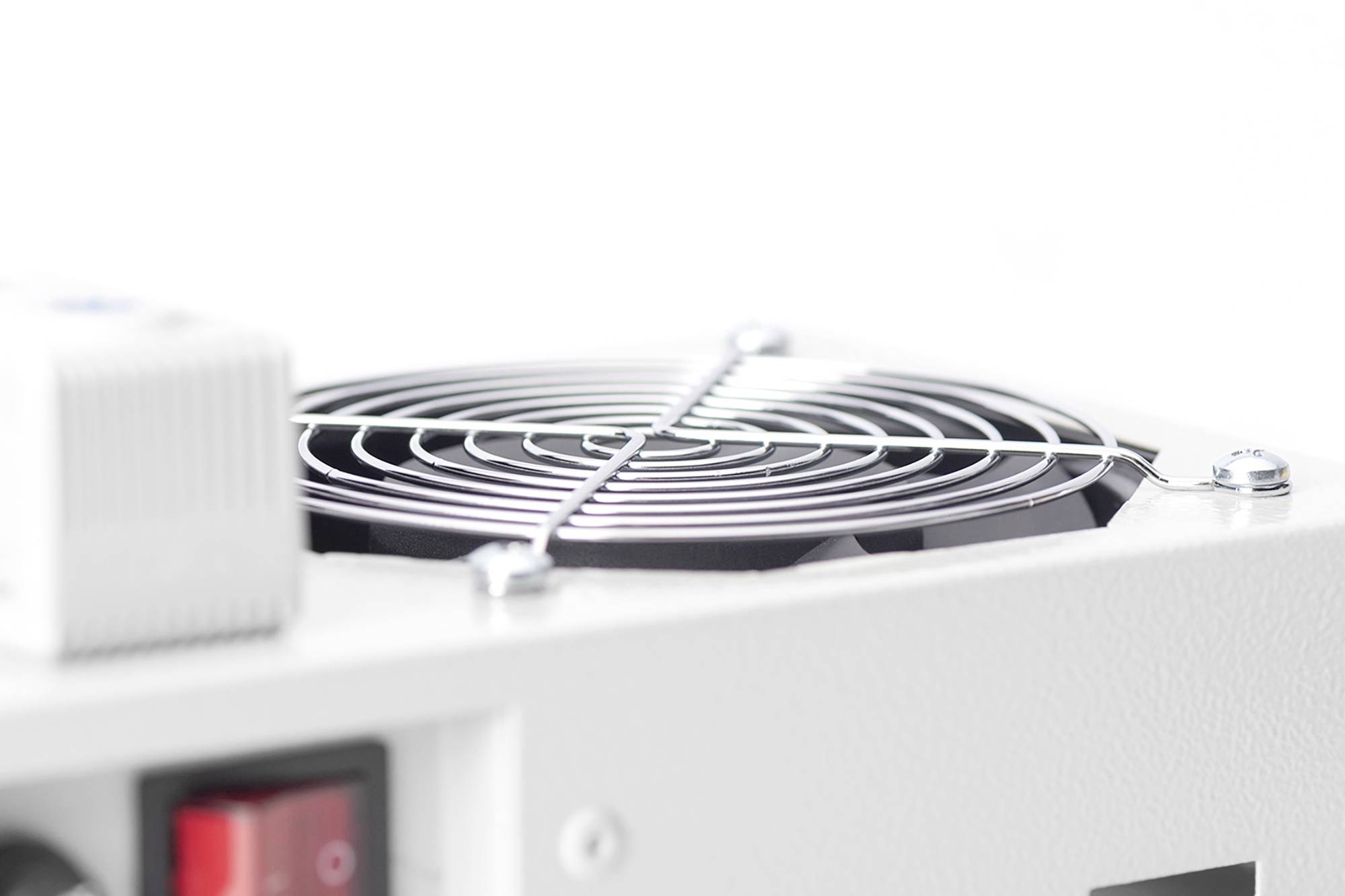 Zbliżenie białego urządzenia elektrycznego z okrągłą kratką wentylacyjną i czerwonym przełącznikiem po lewej stronie.