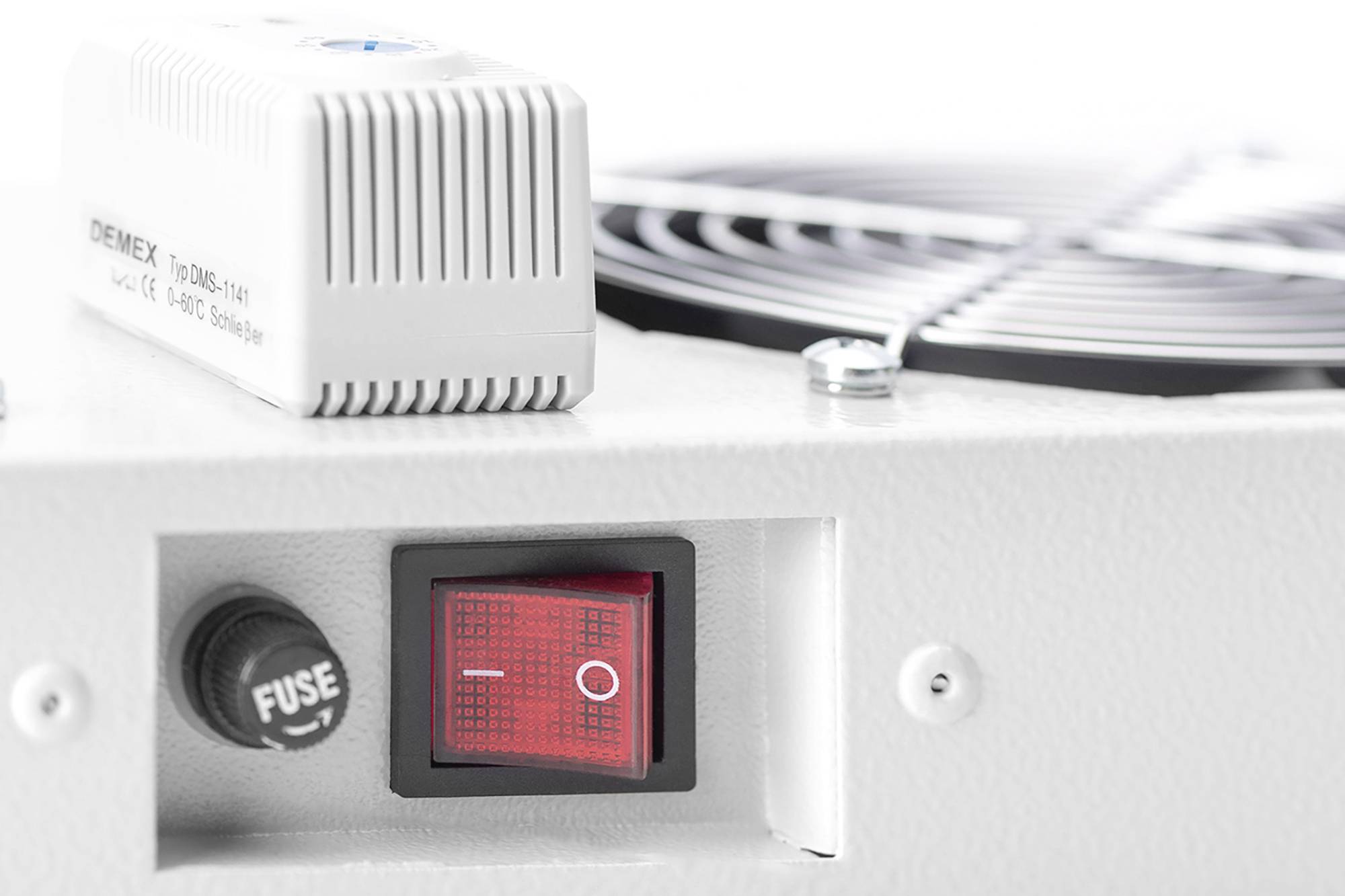 Czerwony przełącznik włączająco-wyłączający na białym urządzeniu, obok którego znajduje się bezpiecznik i kratka wentylatora. Fokus na przełączniku i bezpieczniku.