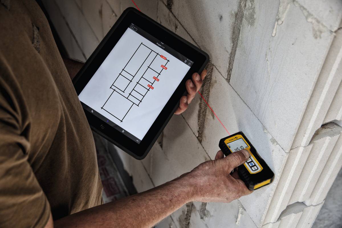 Osoba trzyma tablet z planami budowlanymi i mierzy ścianę za pomocą dalmierza laserowego. Główny nacisk położony jest na precyzyjny pomiar.